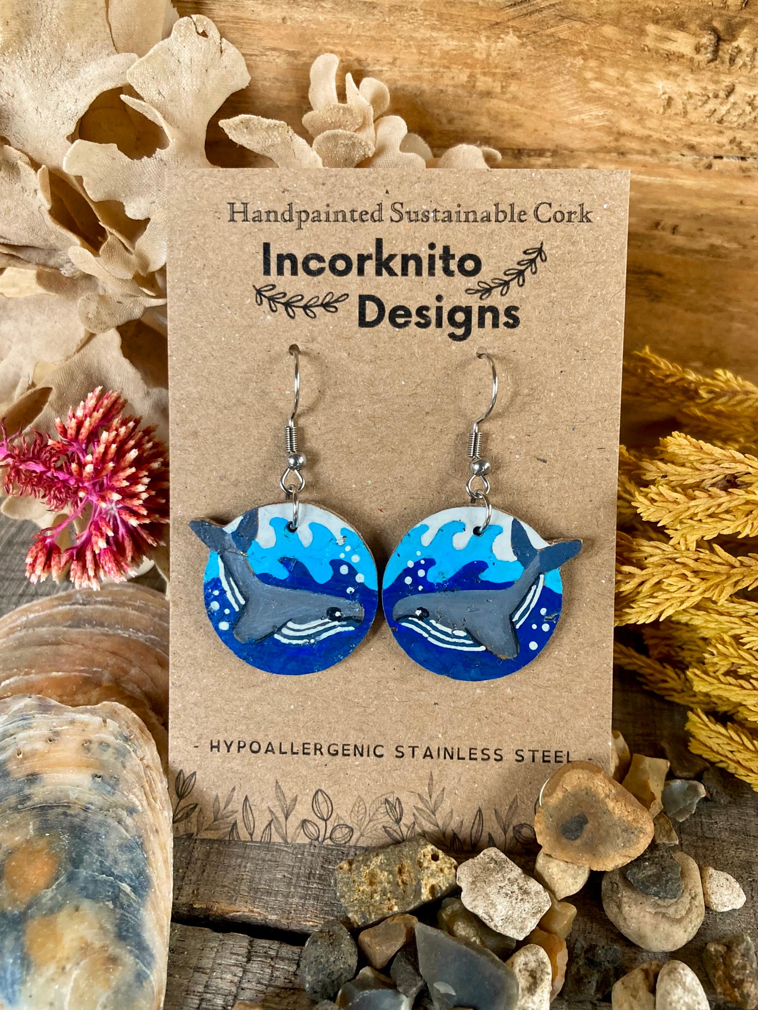 Pair of whale earrings on a 'Incorknito Designs' card with natural elements around.