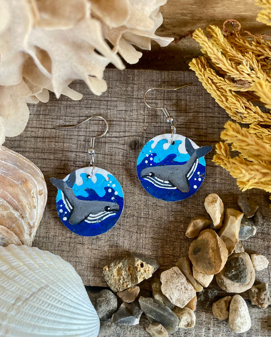 Pair of whale earrings on a wooden surface with shells and stones around