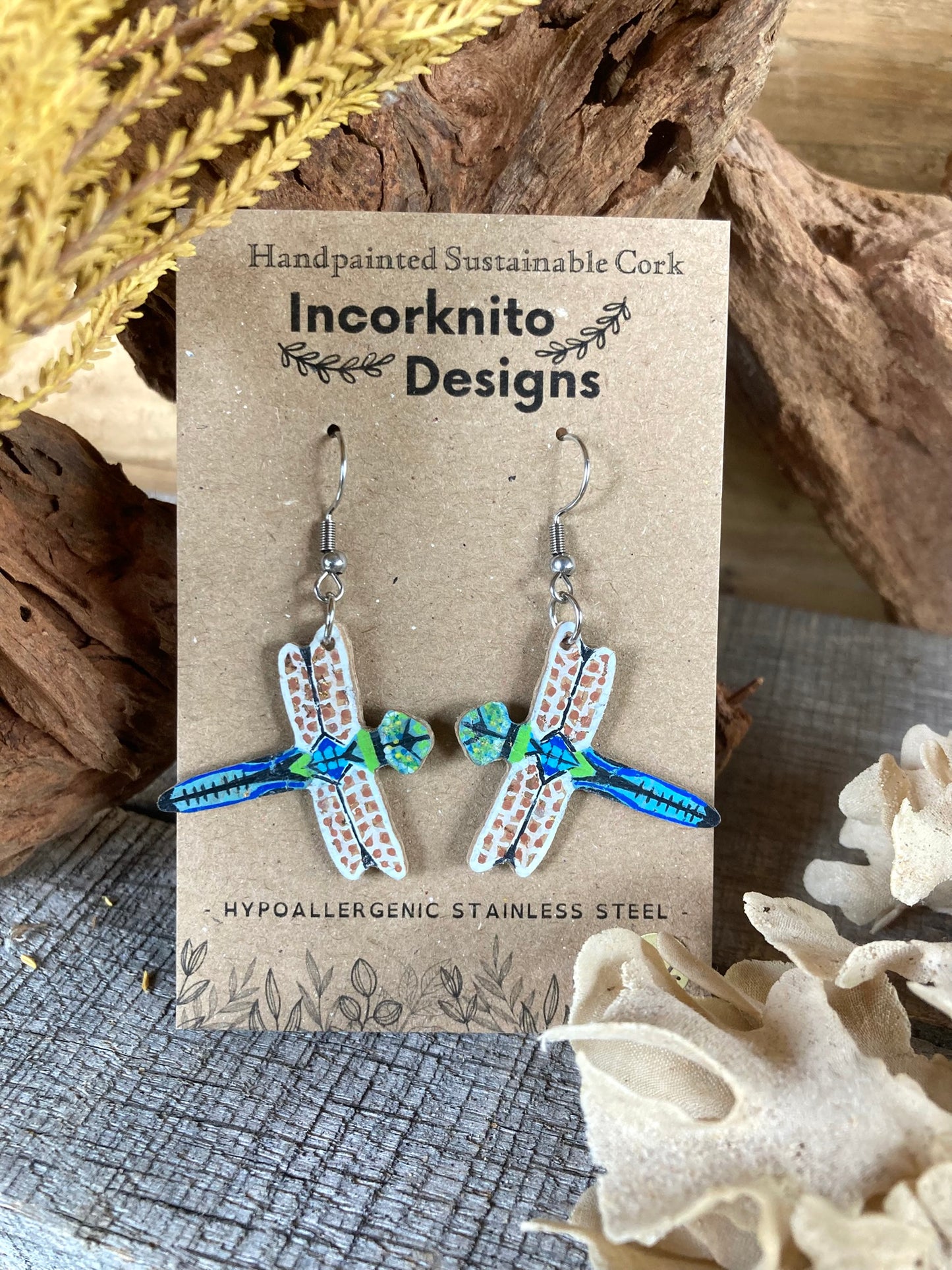 Dragonfly-shaped earrings on a 'Incorknito Designs' card with natural elements in the background.