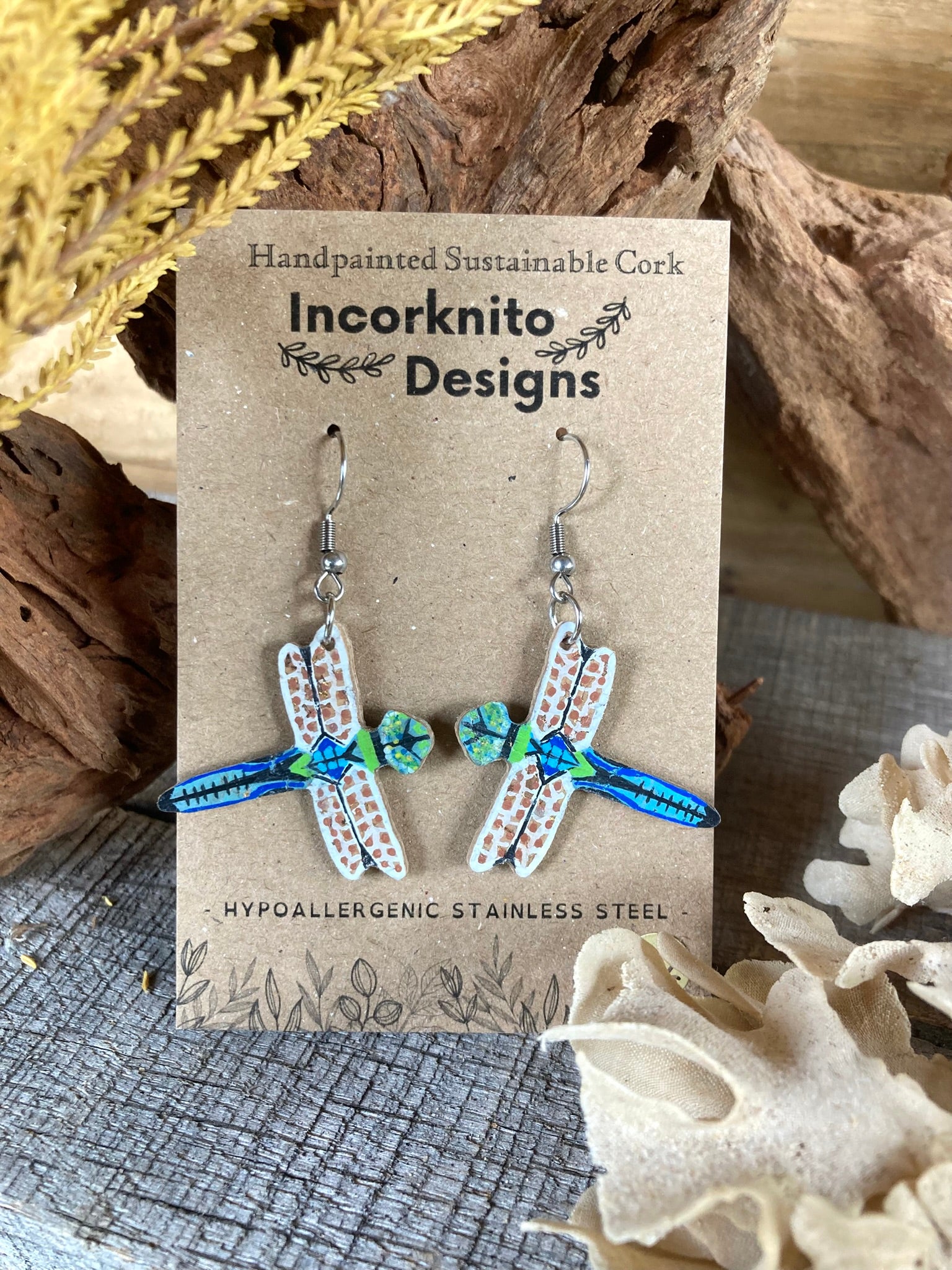 Dragonfly-shaped earrings on a 'Incorknito Designs' card with natural elements in the background.
