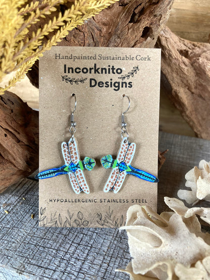 Dragonfly-shaped earrings on a 'Incorknito Designs' card with natural elements in the background.