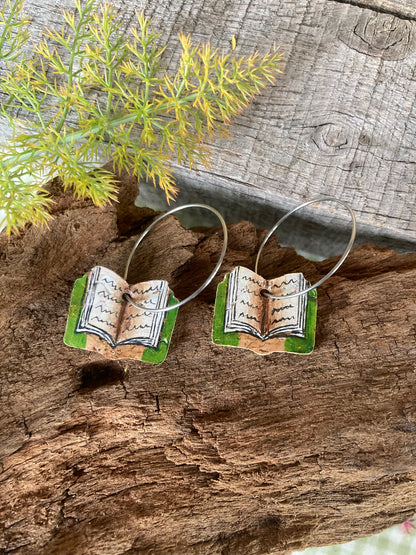 Open book-shaped earrings on a wooden surface with greenery