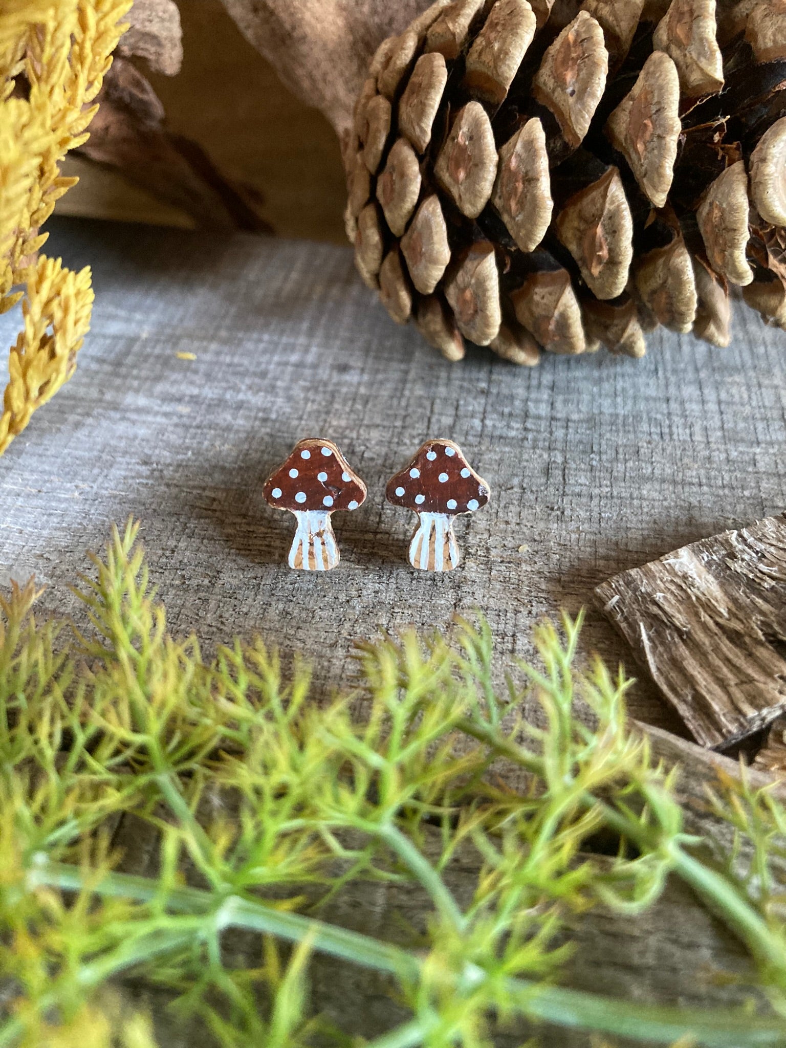 Pair of brown mushroom-shaped earrings on a textured surface with pine cones and greenery.