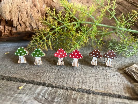 Set of mushroom-shaped earrings in red, green and brown with white polka dots on a wooden surface with greenery