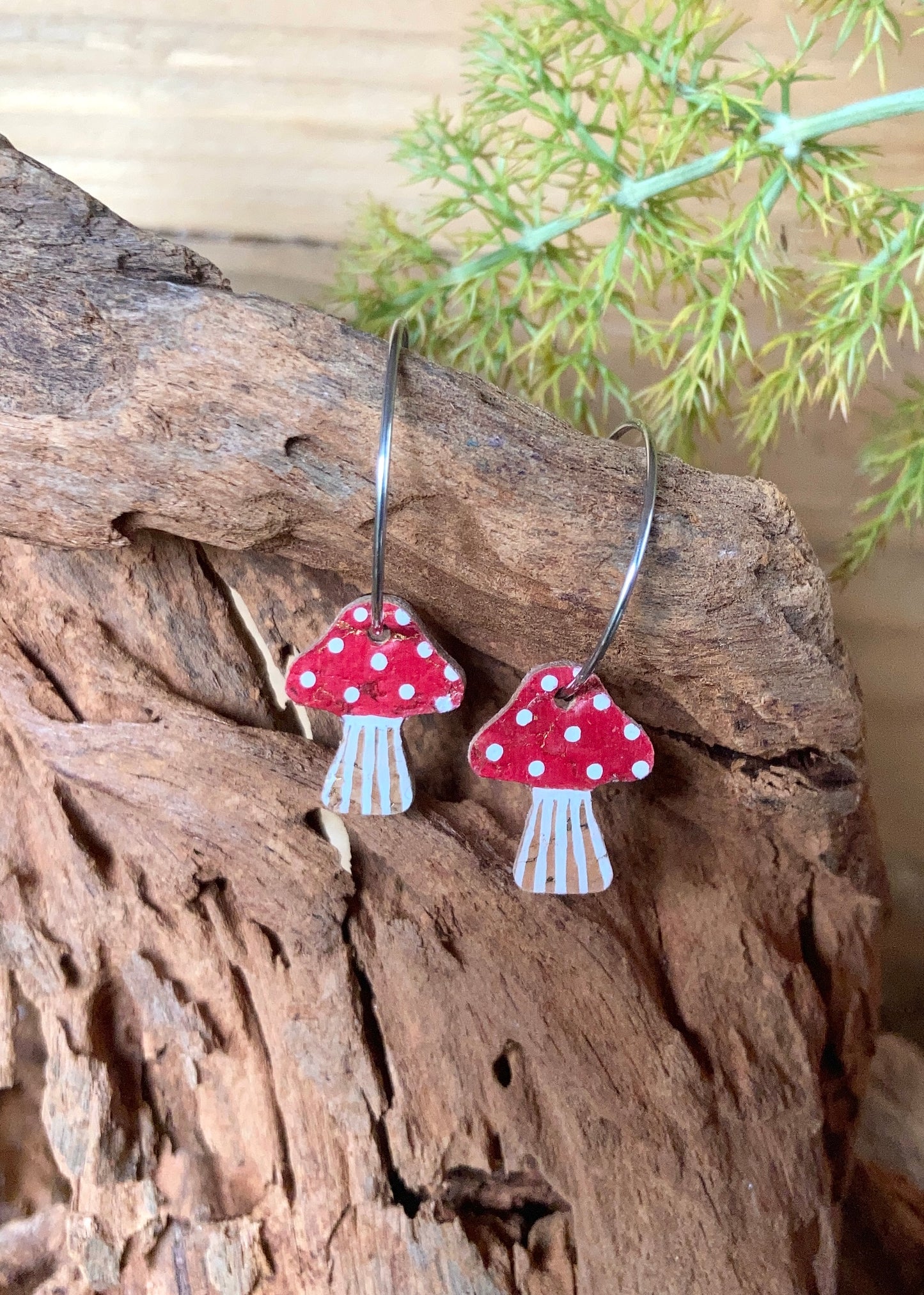 Red and white mushroom-shaped earrings hanging on a wooden surface with greenery in the background.