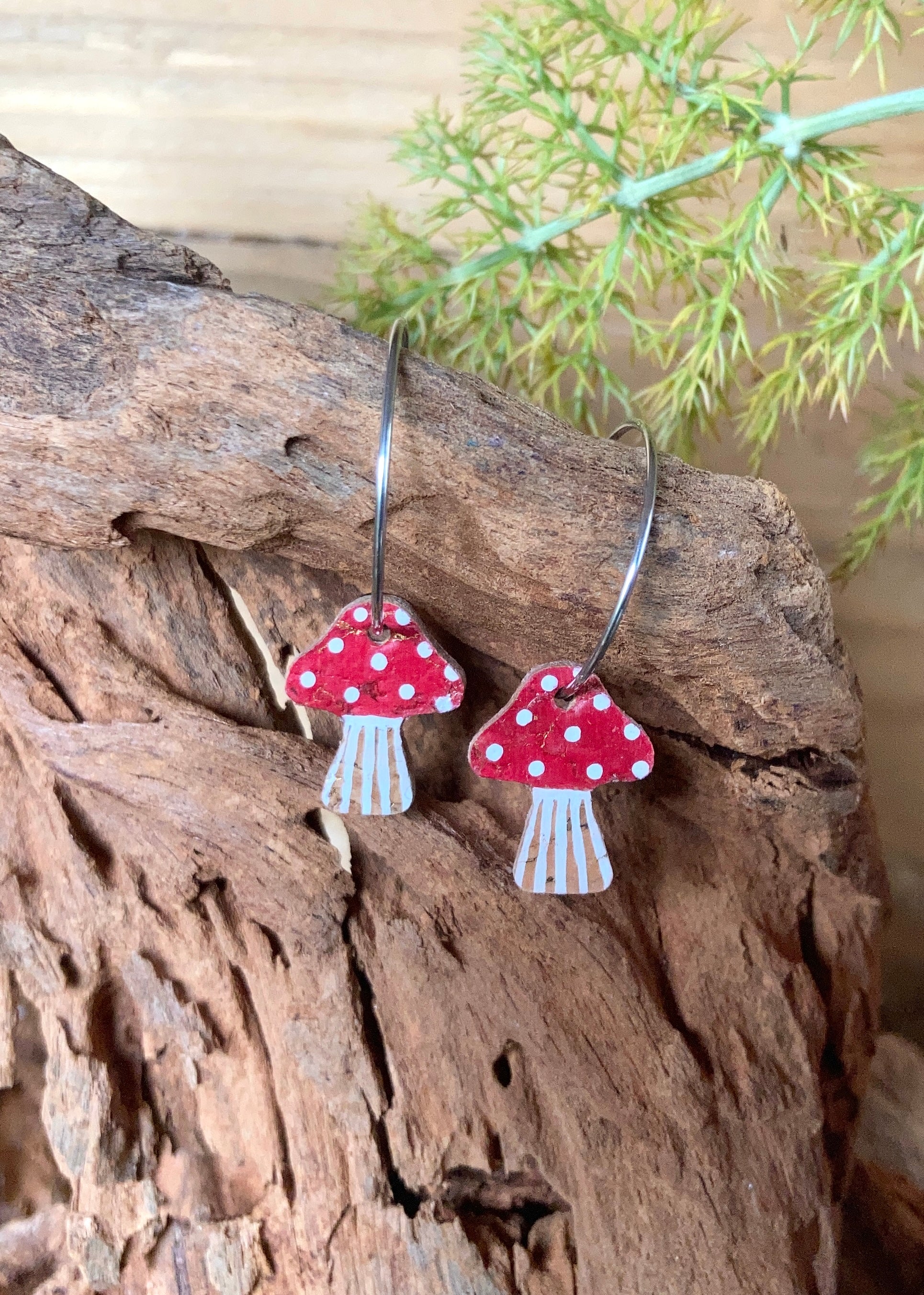 Red and white mushroom-shaped earrings hanging on a wooden surface with greenery in the background.