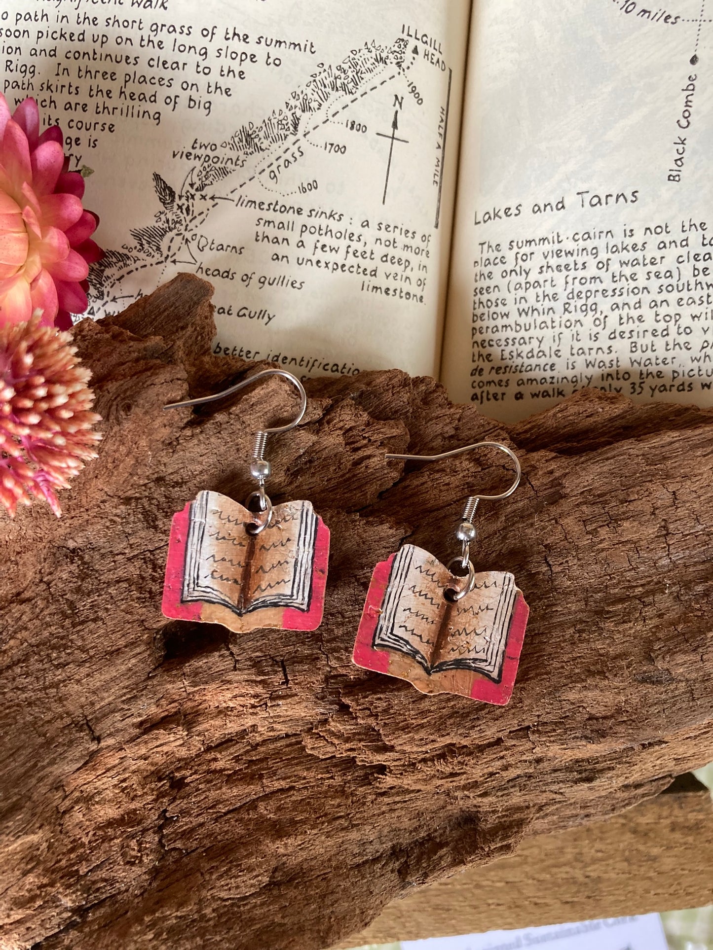 Book-themed earrings on a wooden surface with an open book in the background