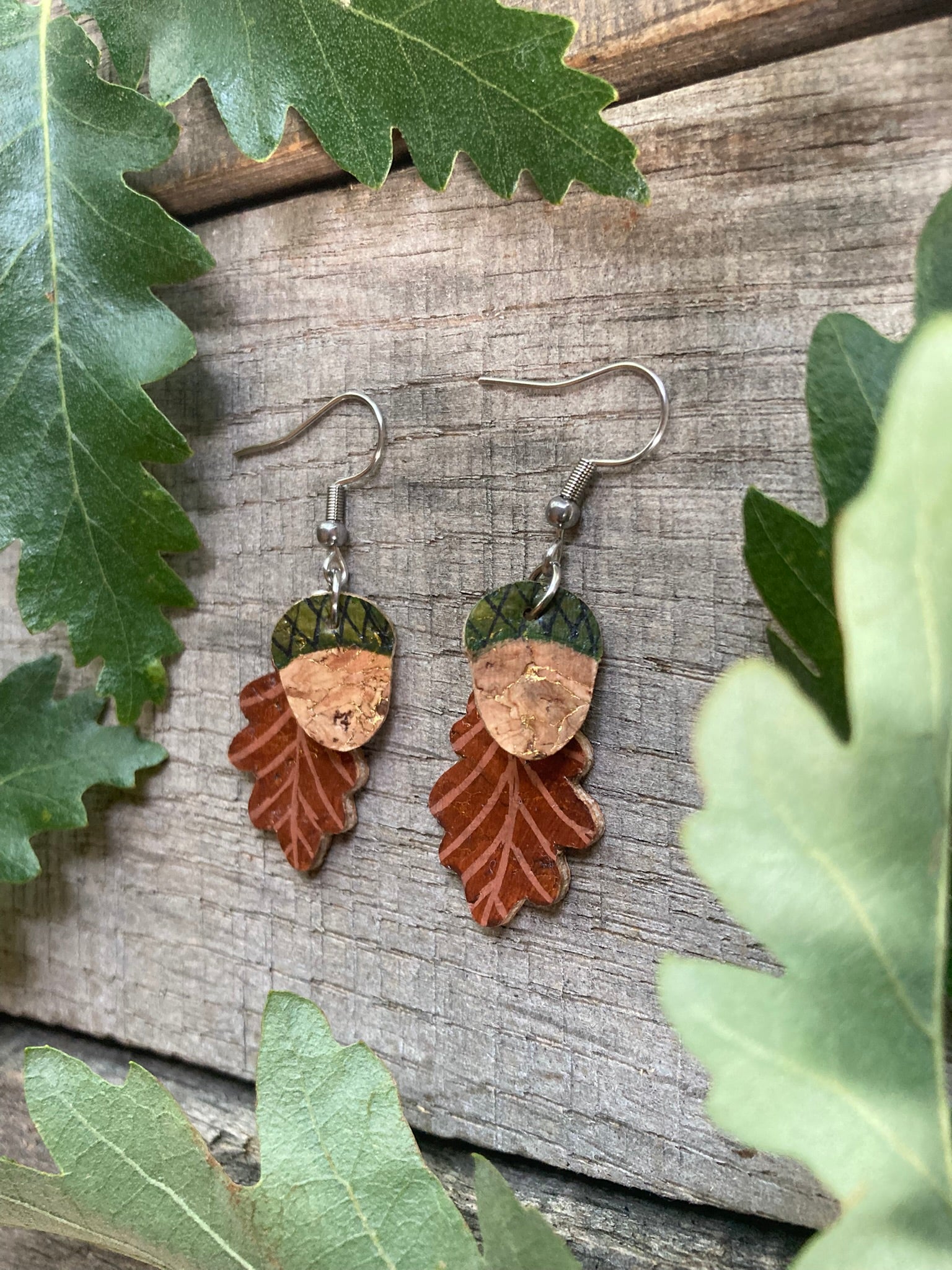 Pair of leaf-shaped earrings on a wooden surface with green leaves around.