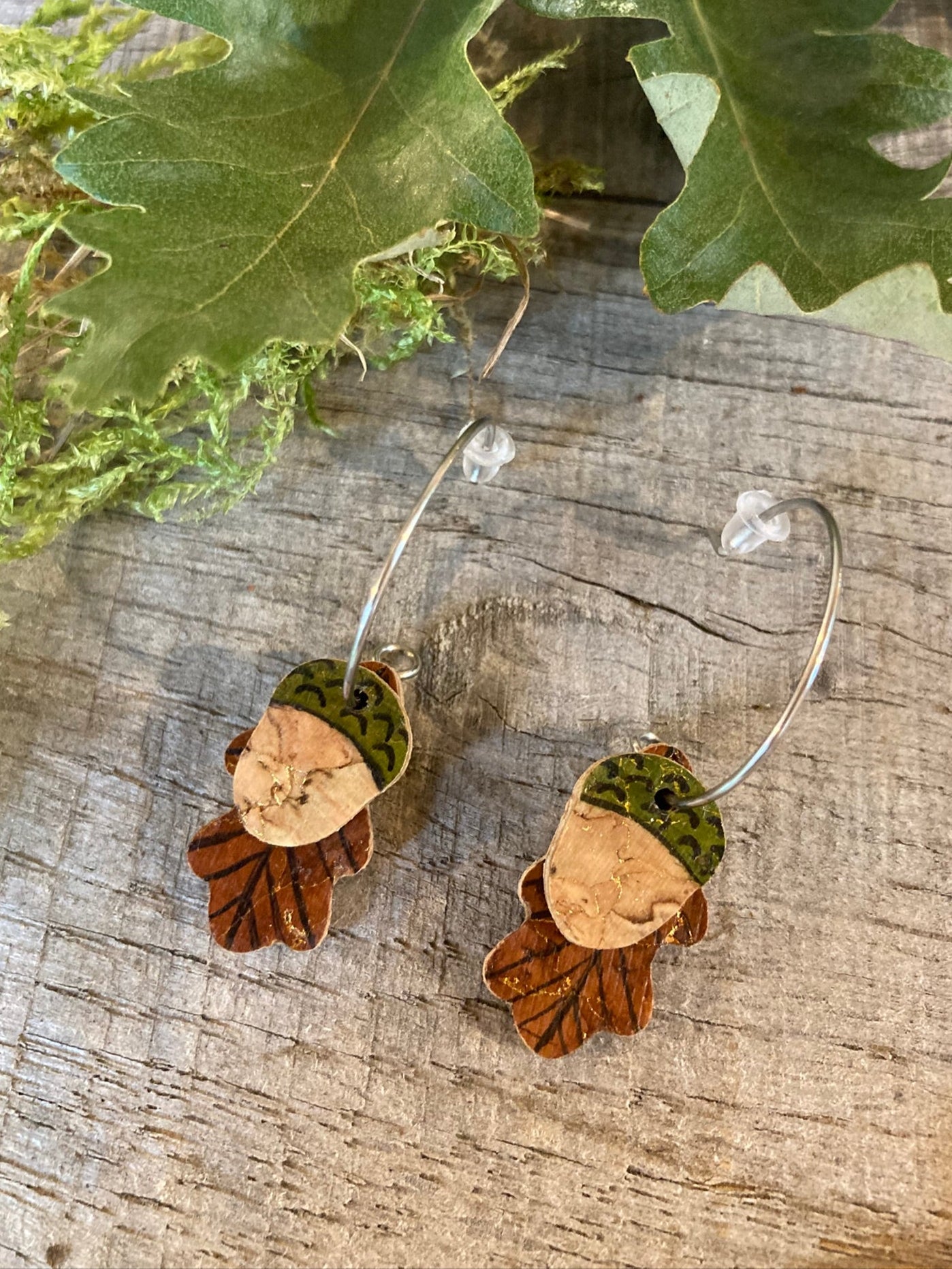 Pair of leaf-shaped earrings on a wooden surface with green leaves.