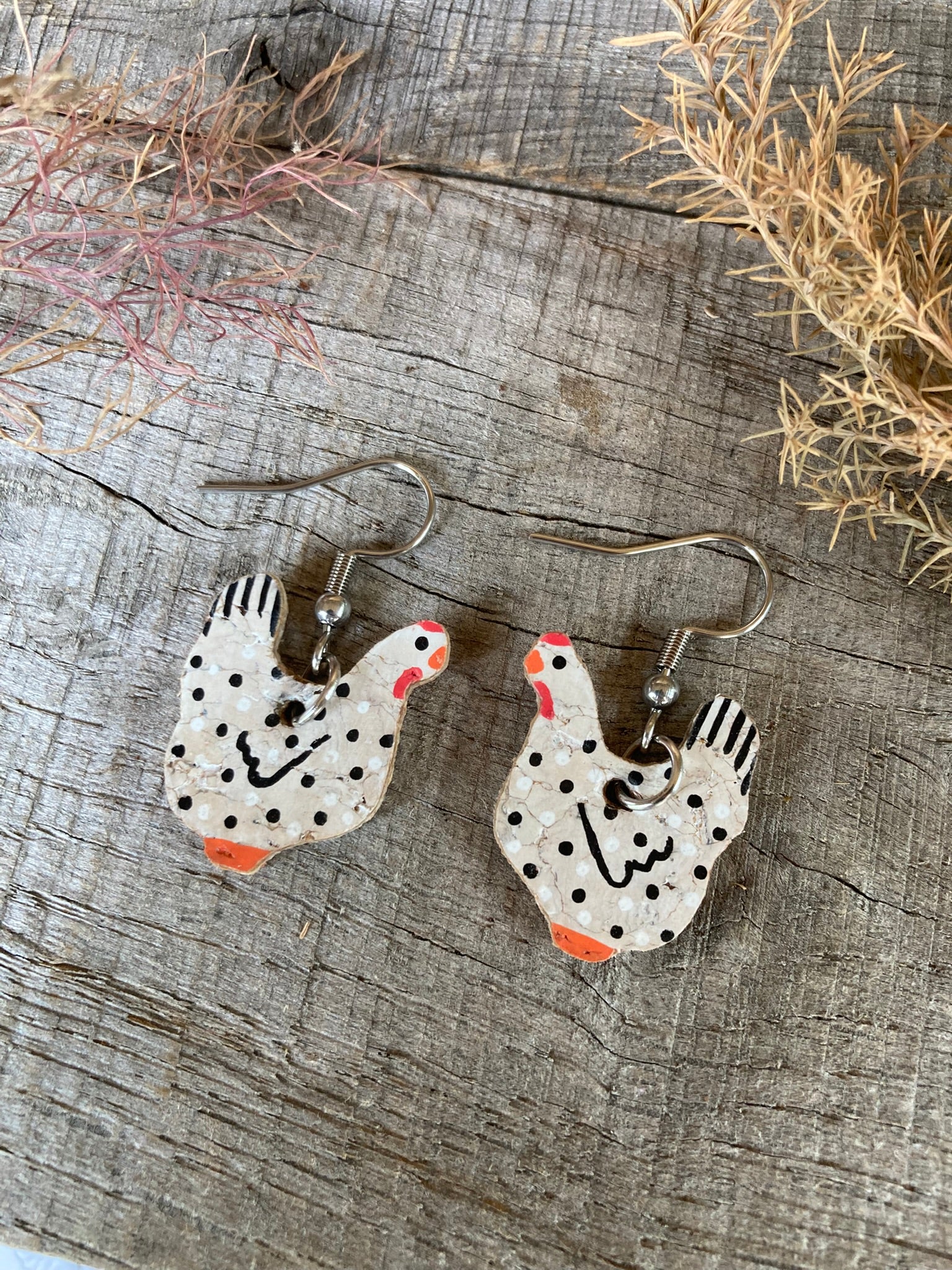 Pair of chicken-shaped earrings on a wooden surface with dried plants.