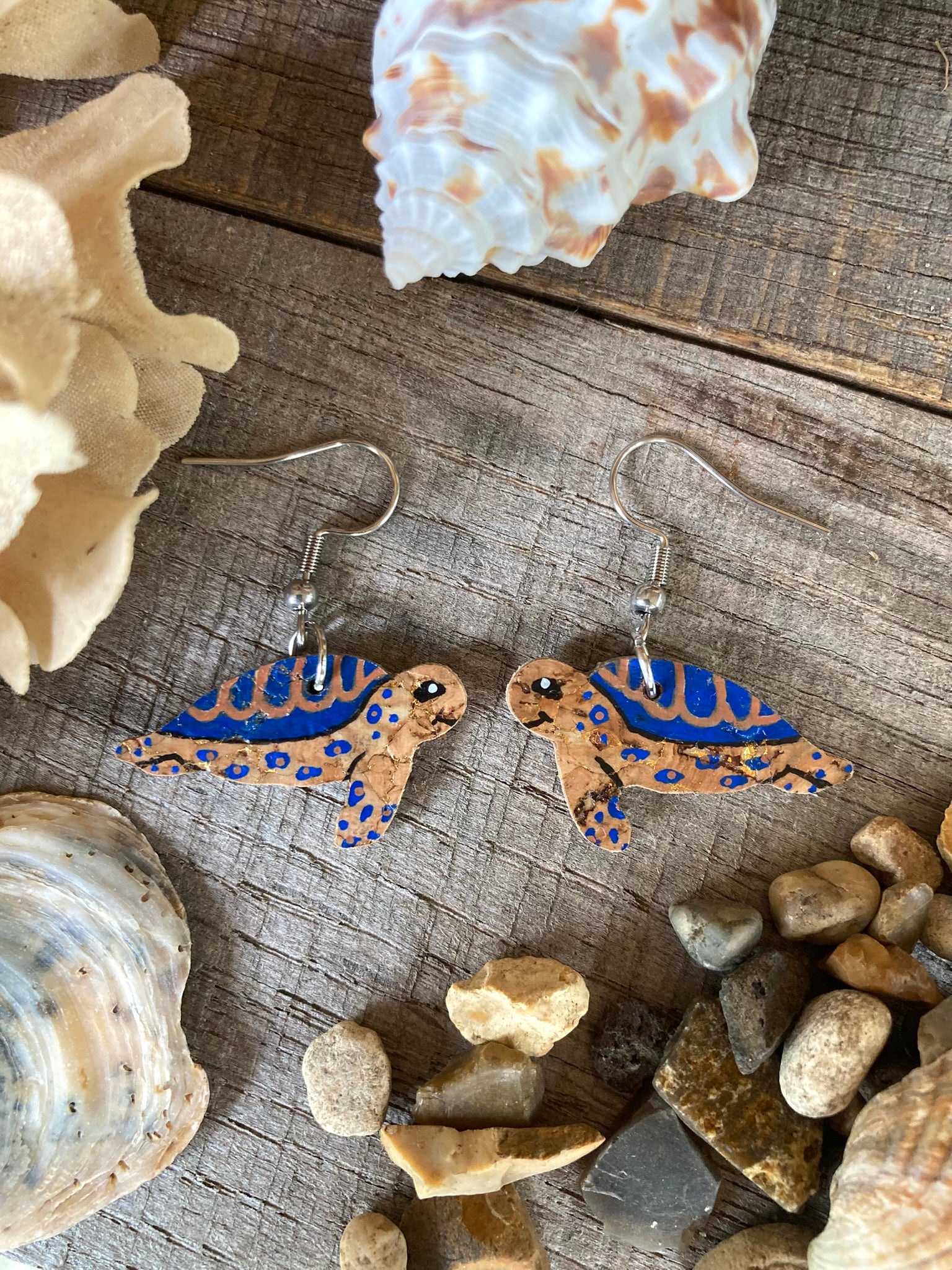 Turtle-shaped earrings with blue accents on a wooden surface with seashells and stones.