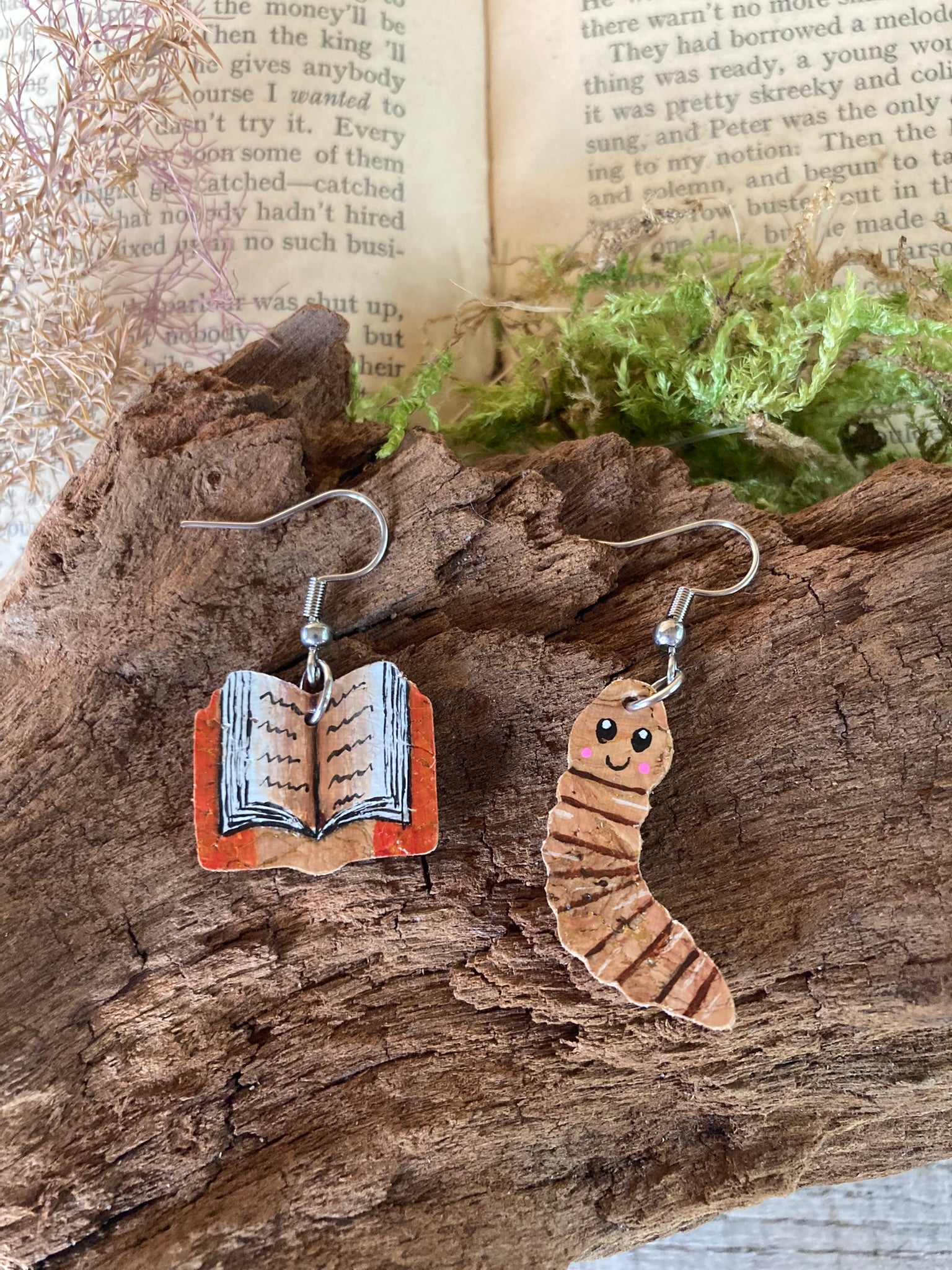 Pair of earrings shaped like a book and a worm (=bookworm) on a wooden surface with a book in the background.