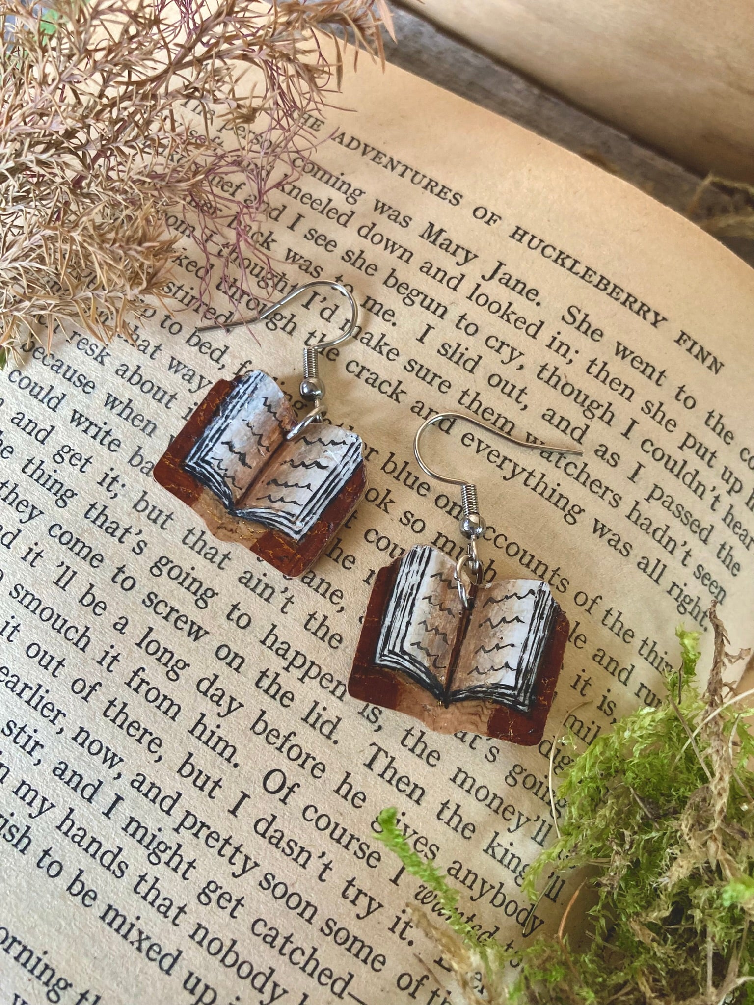 Book-themed earrings on an open book with natural elements.