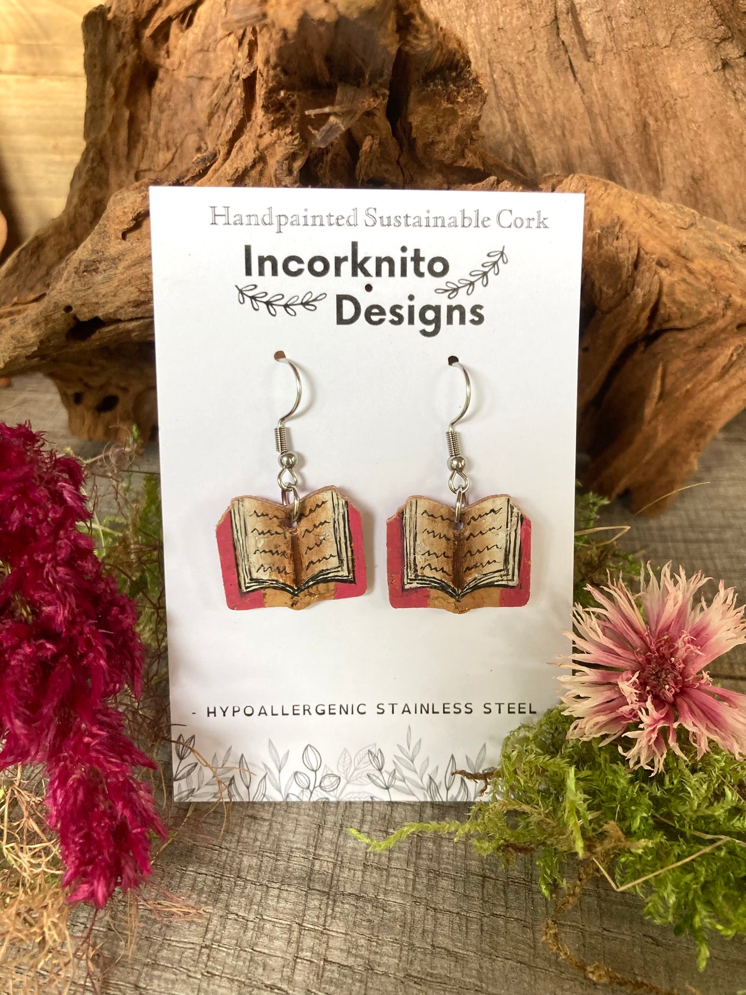Book-shaped earrings on a card with 'Incorknito Designs' branding, surrounded by natural elements.