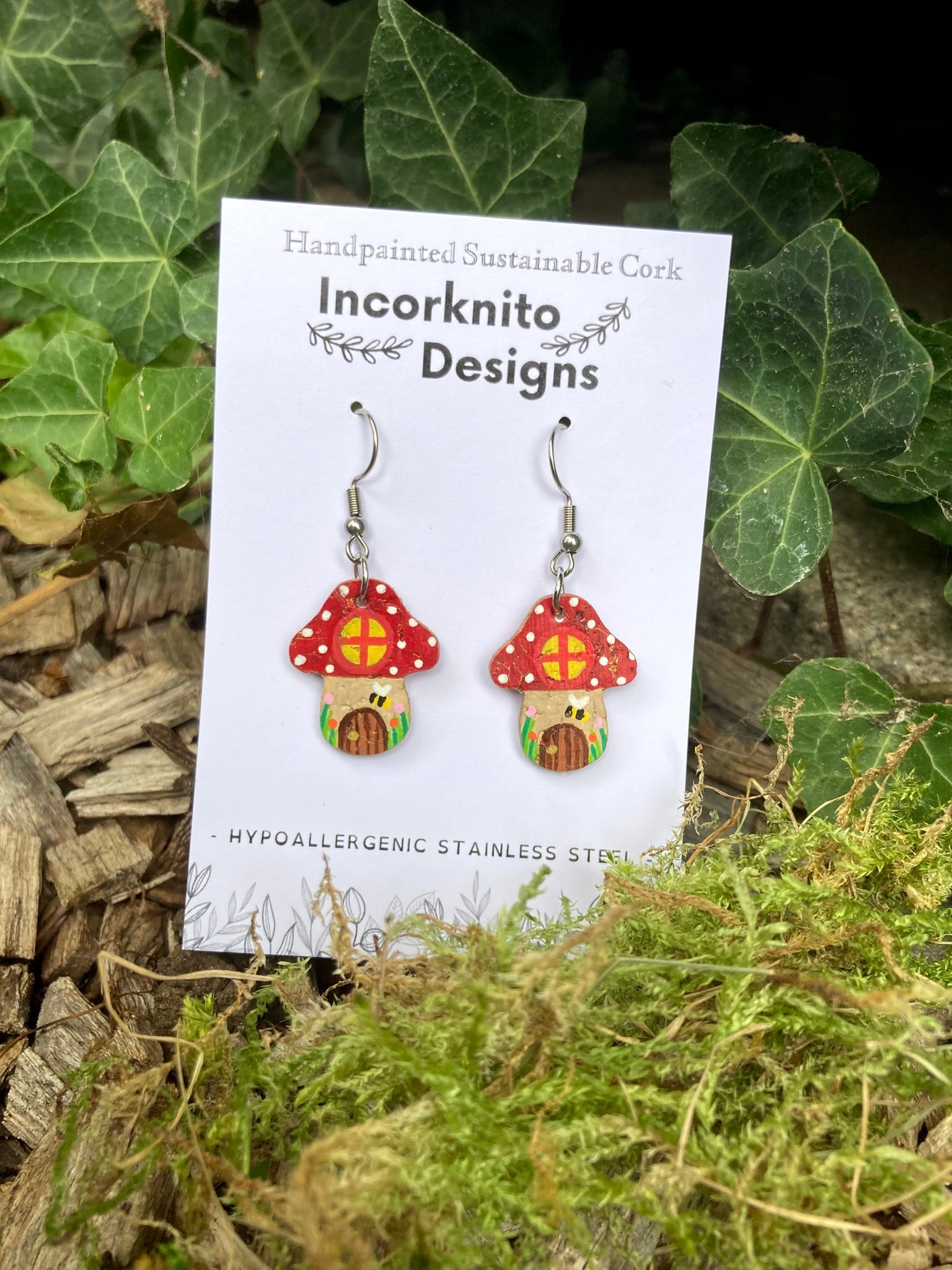 Incorknito Designs mushroom-shaped earrings on a card with greenery in the background