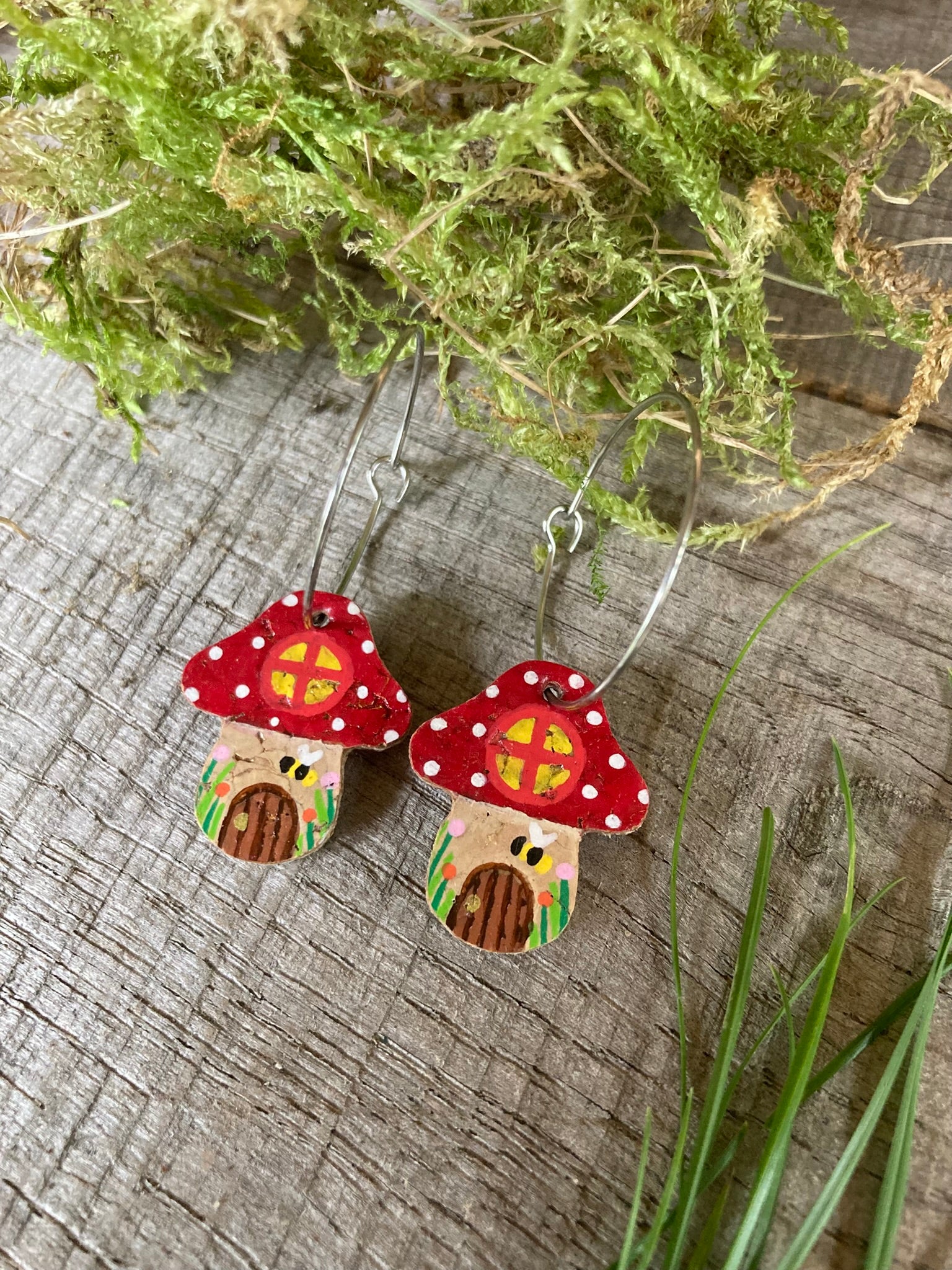 Mushroom-shaped earrings with red caps and brown stems on a wooden surface with greenery.