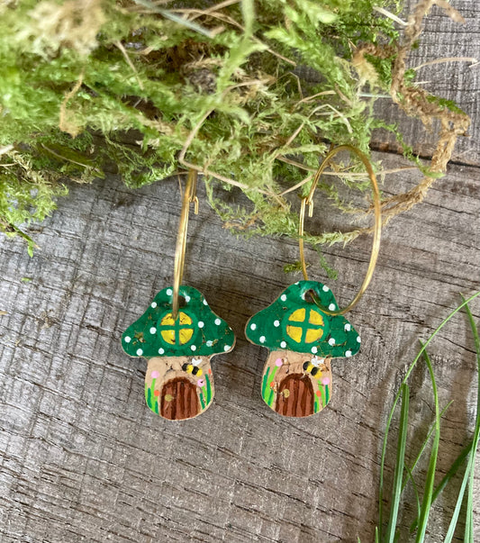 Fairy house toadstool earrings featuring a little bee and wooden door with flowers around photographed on a wooden surface with greenery