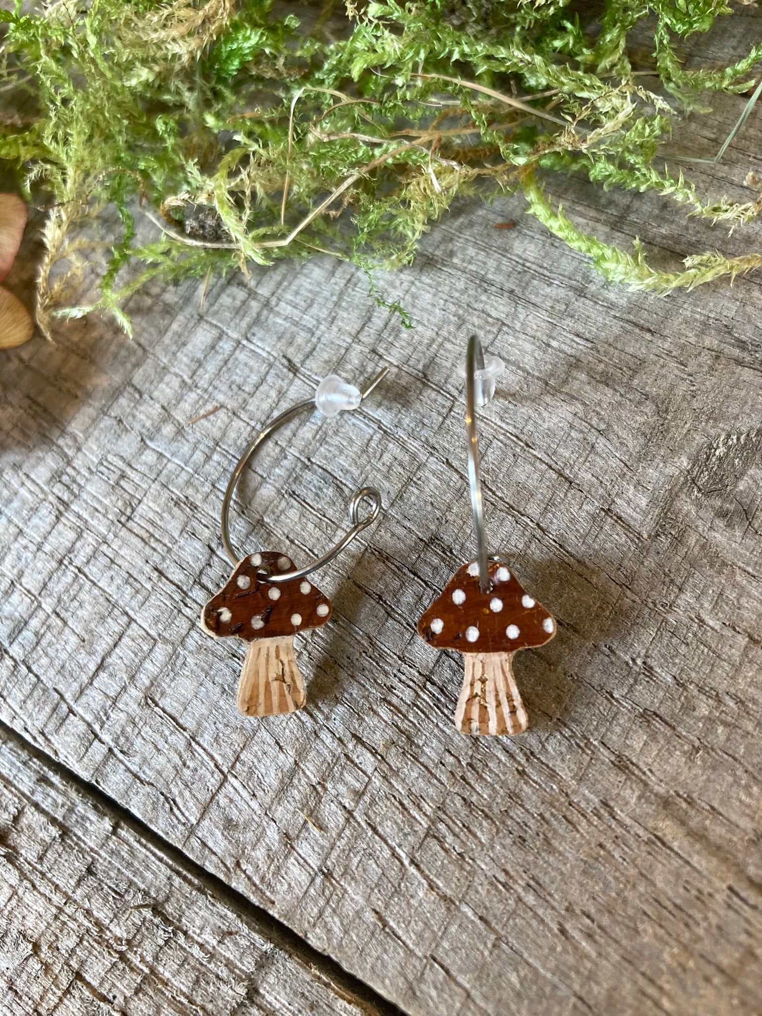 brown spotted toadstool-shaped earrings on a wooden surface with greenery in the background