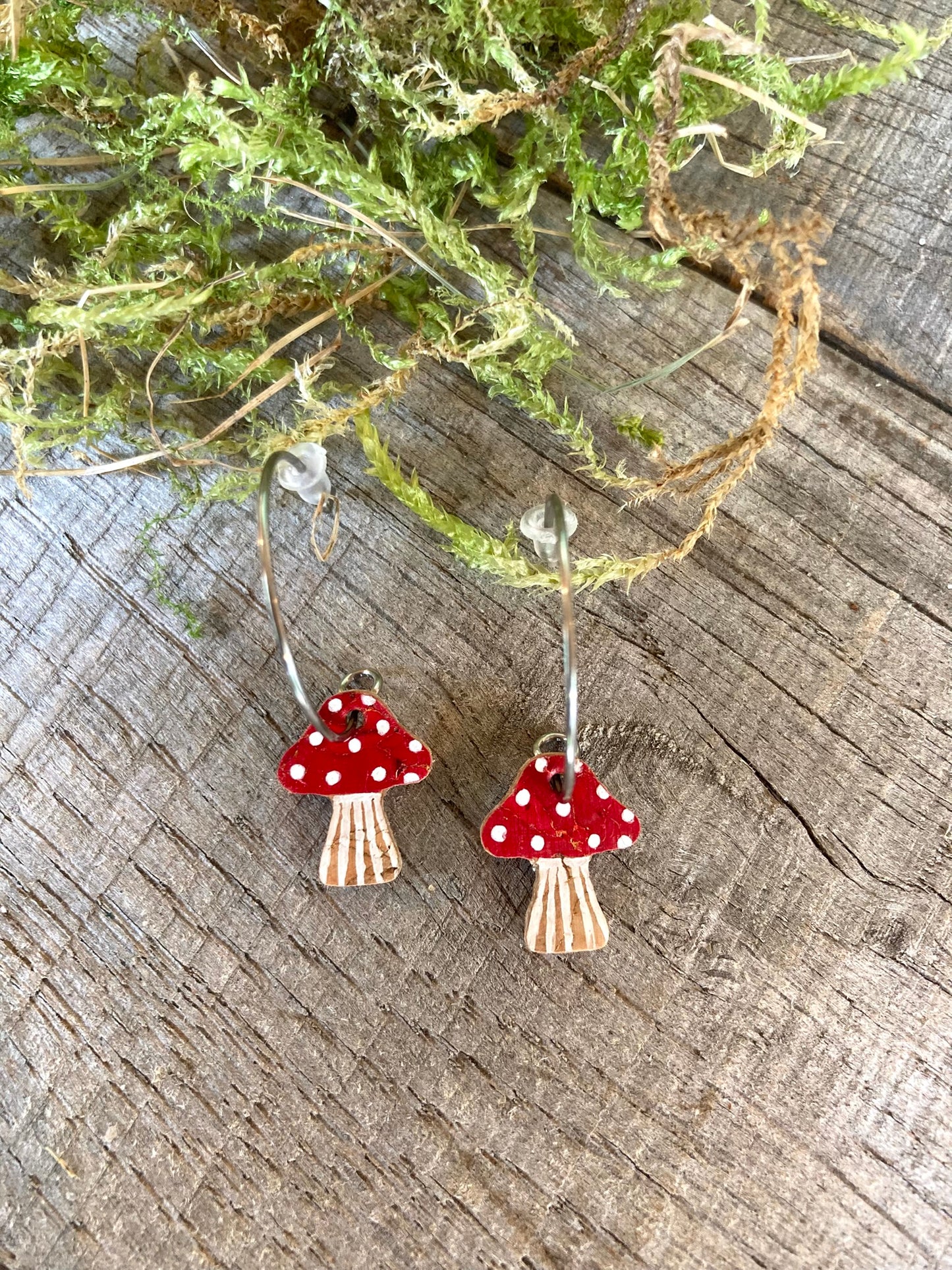 Mushroom-shaped earrings with red caps and white spots on a wooden surface with greenery.