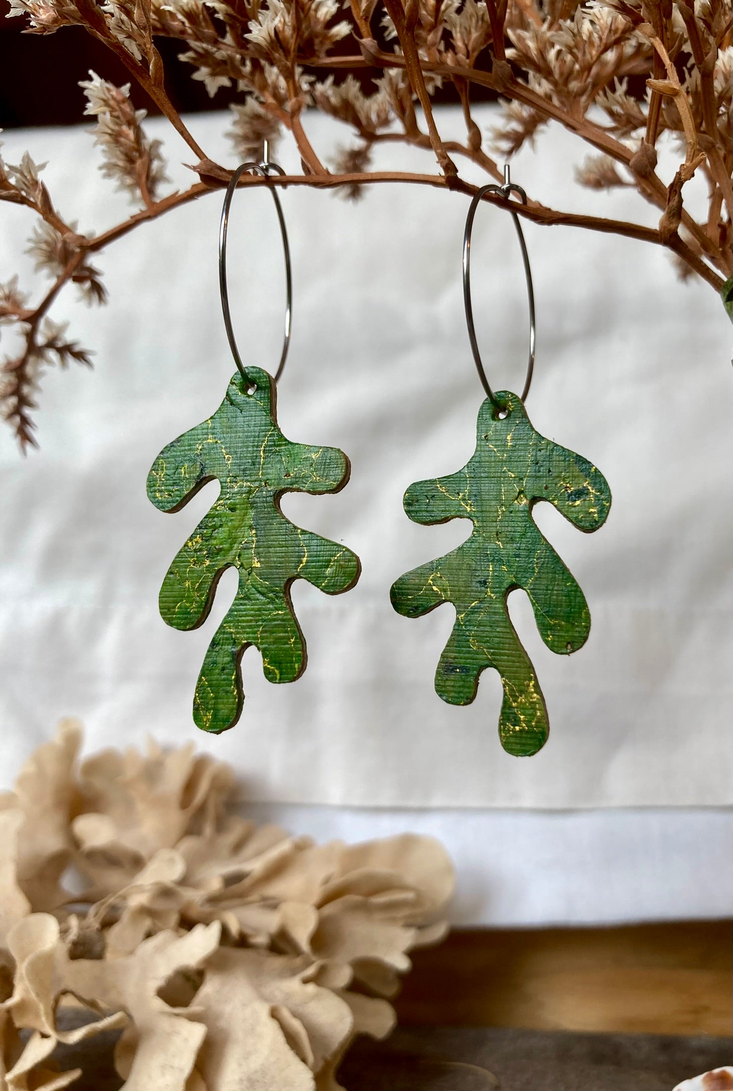 Green leaf-shaped earrings hanging against a neutral background with dried plants.