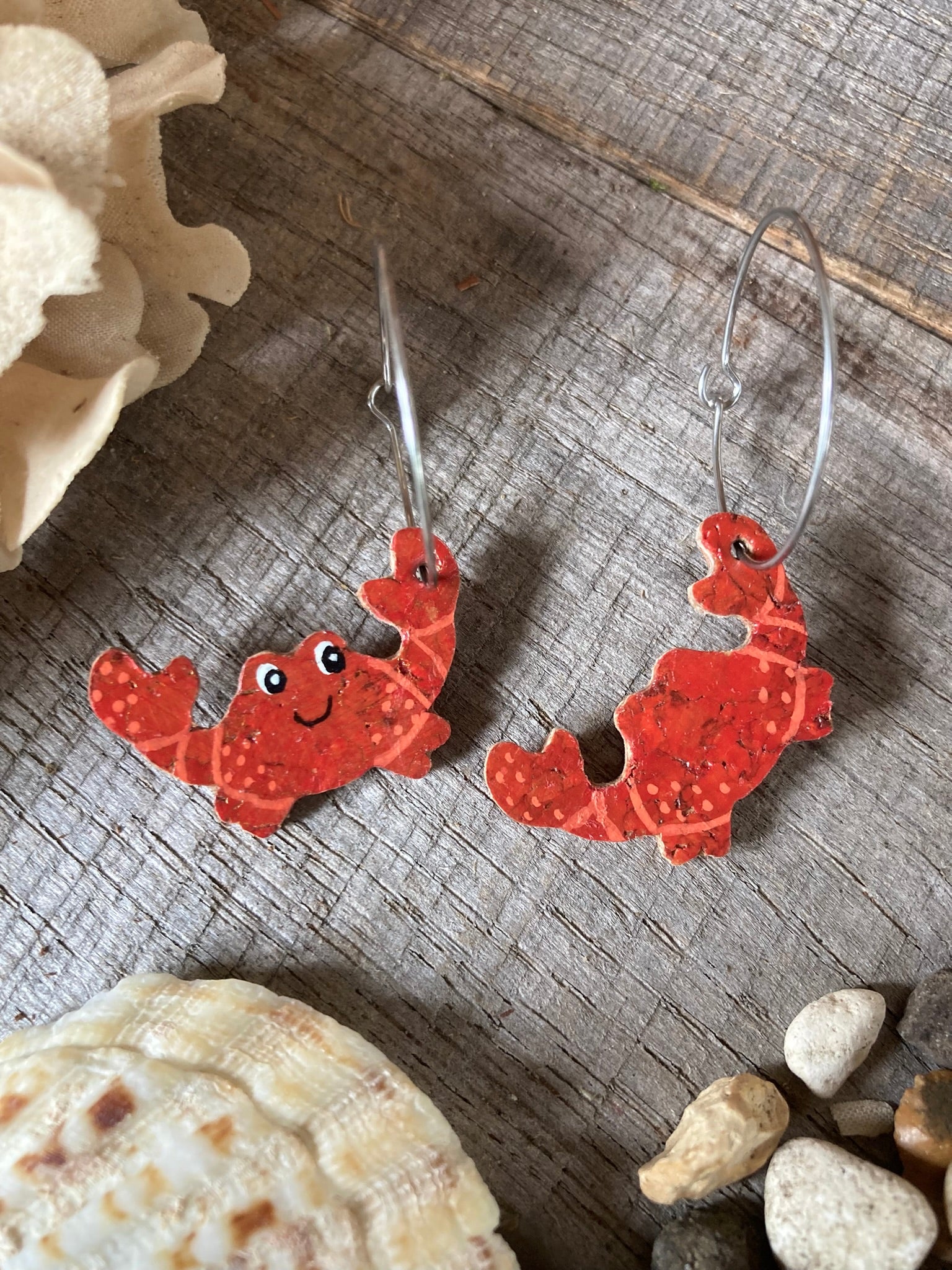 Red crab-shaped earrings on a wooden surface with seashells and stones.