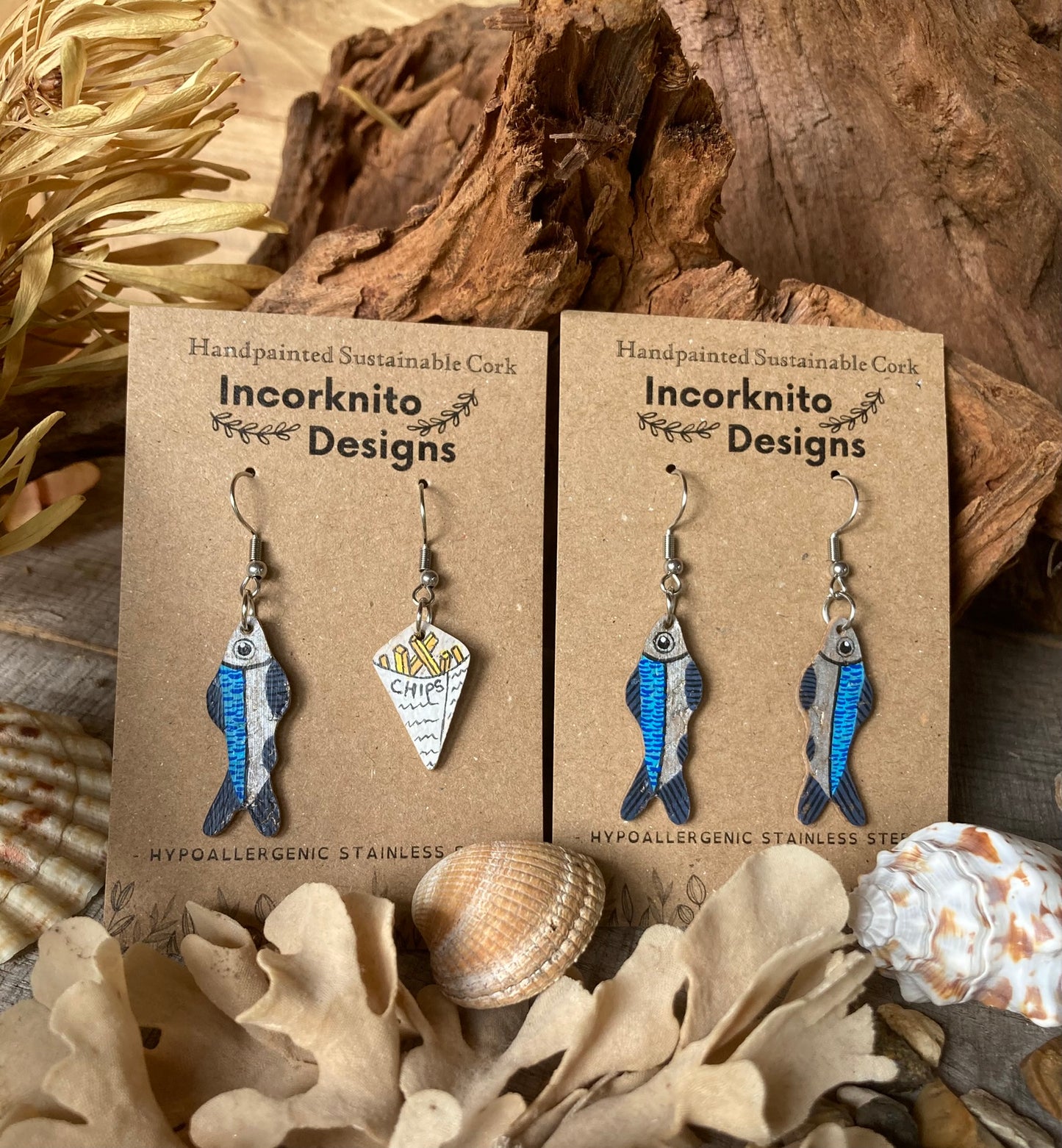 Pair of fish-shaped earrings and fish and chip painted earrings on Incorknito Designs cards with natural elements in the background