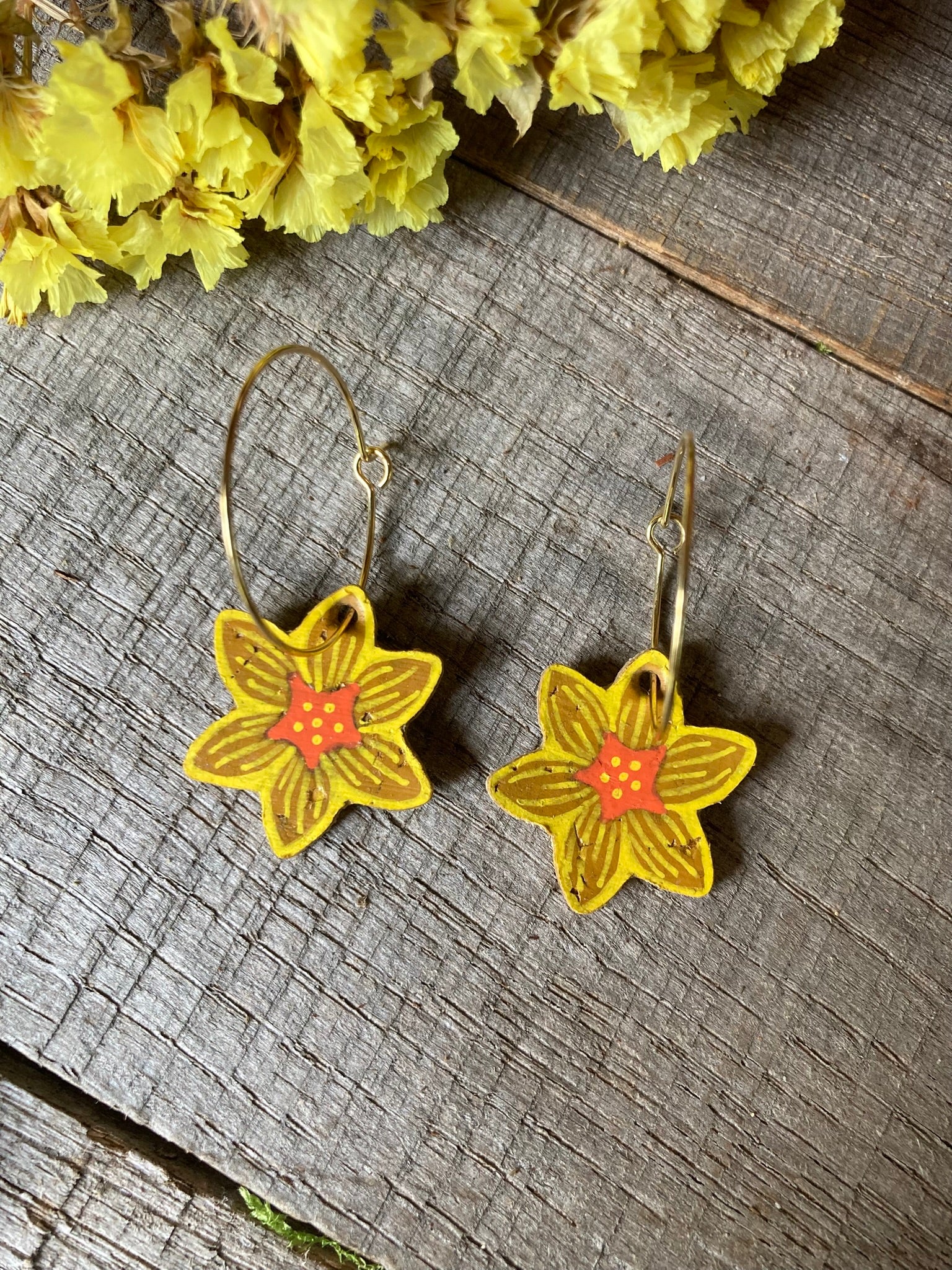 Daffodil shaped earrings on a wooden surface with yellow flowers in the background