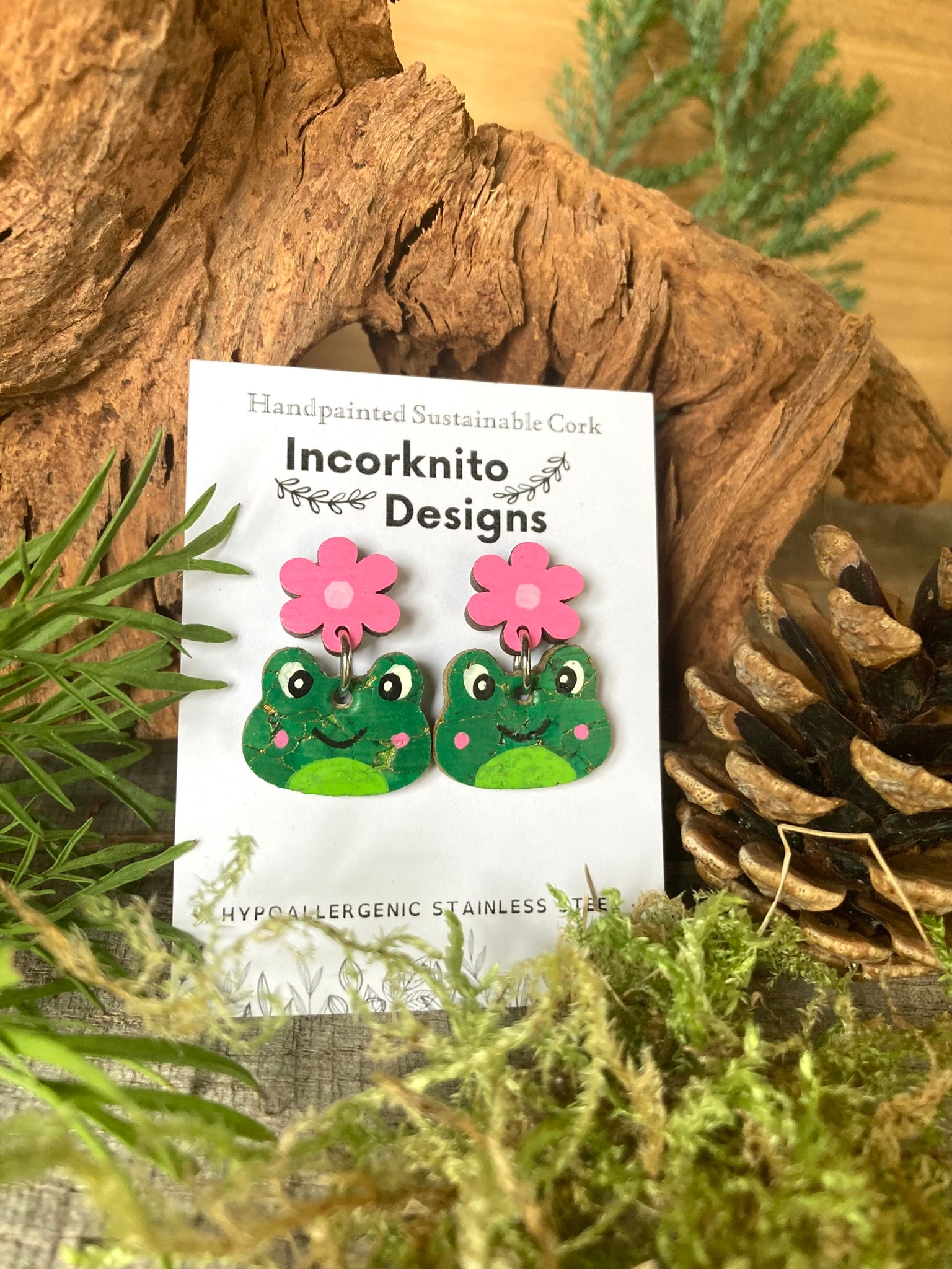 Frog-shaped earrings with pink flowers on a 'Incorknito Designs' card, surrounded by natural elements.