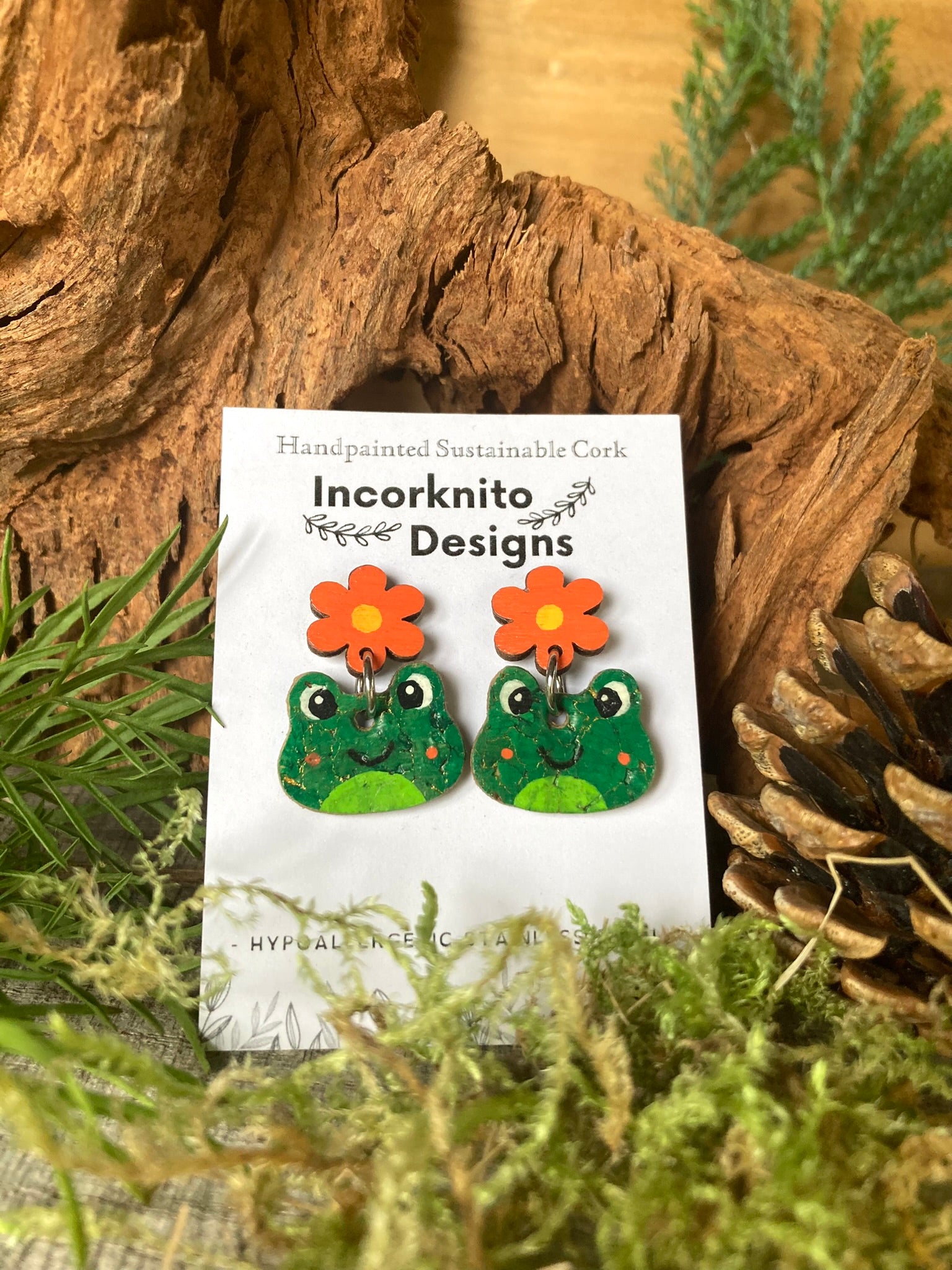 Pair of green frog-shaped earrings with orange flowers on a white card labeled 'Incorknito Designs' against a natural wood and foliage background.