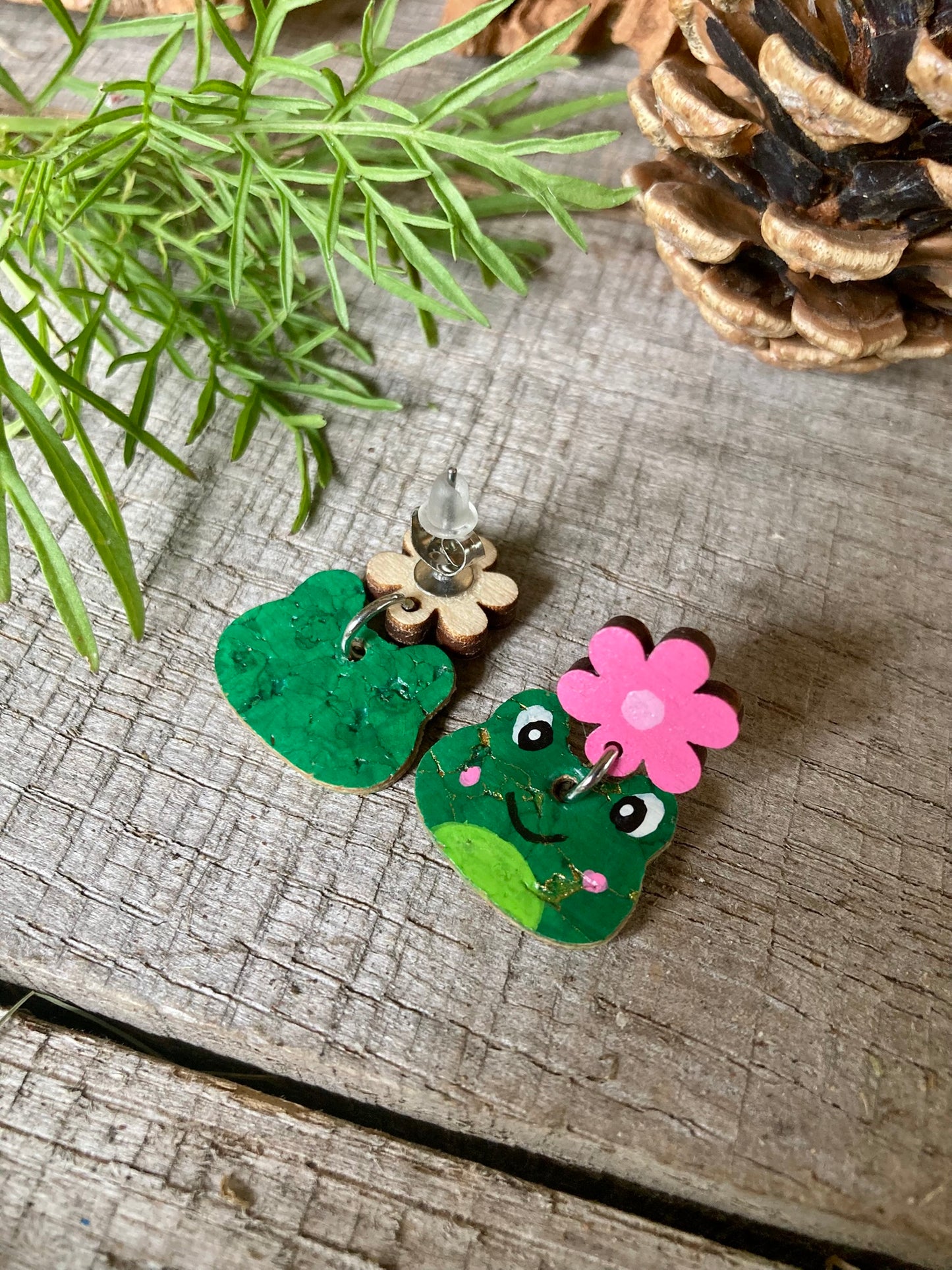 Pair of green frog earrings with pink flower on a wooden surface with foliage and pine cone.