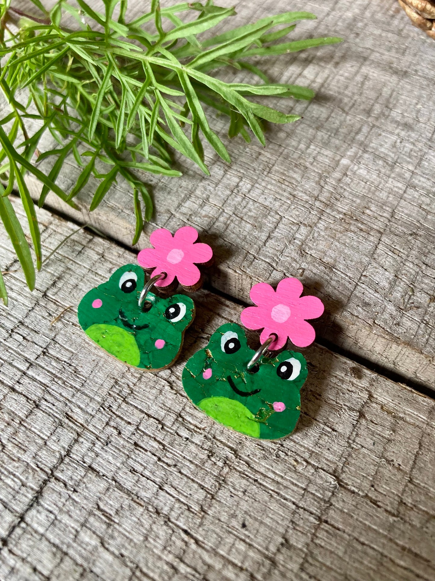 Frog-shaped earrings with pink flowers on a wooden surface with green leaves.