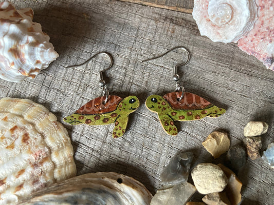 Green turtle handpainted cork earrings on a wooden background with natural seaside elements around