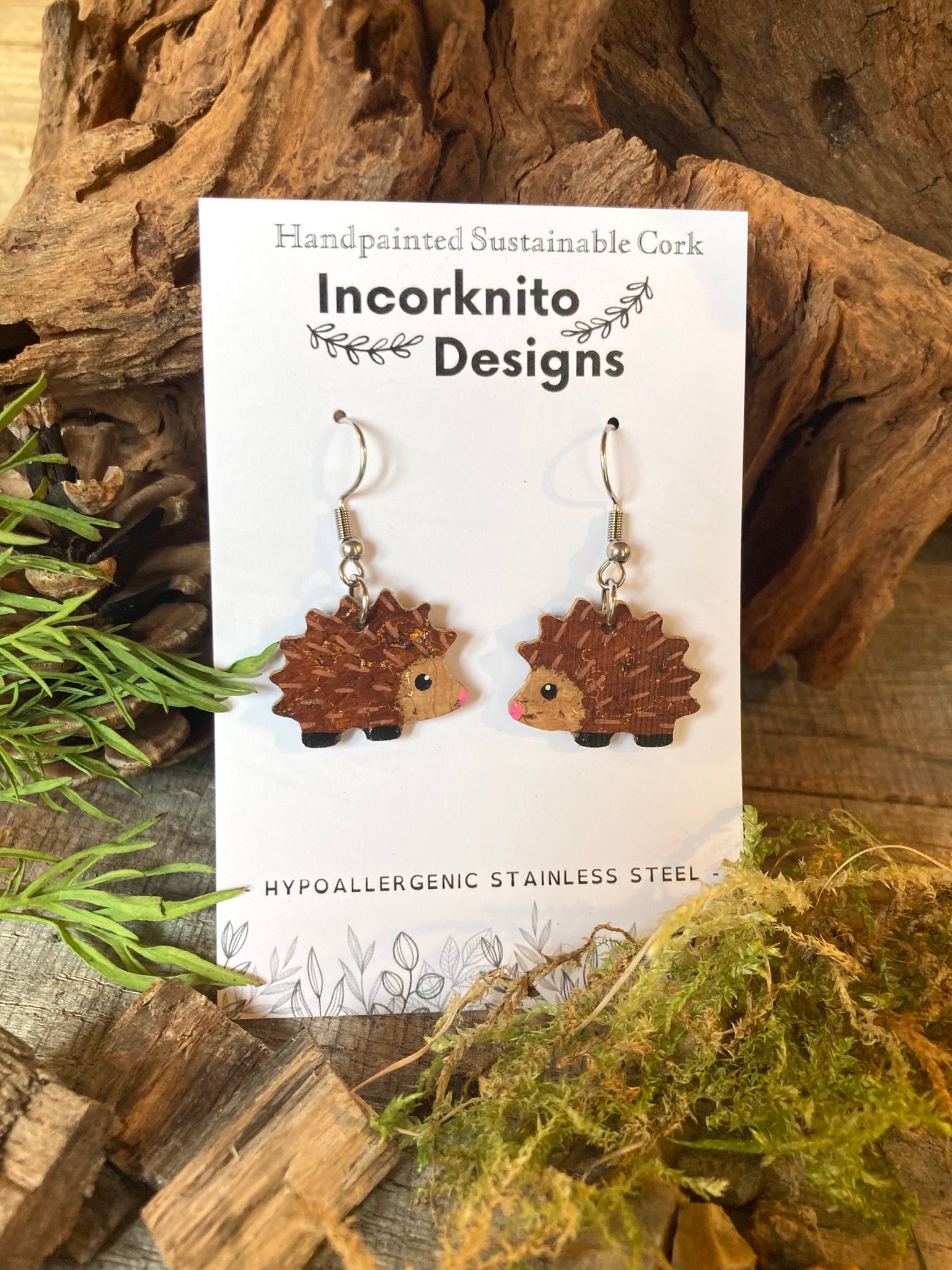 Hedgehog-shaped earrings on a card with 'Incorknito Designs' branding, set against a natural wood and moss background.