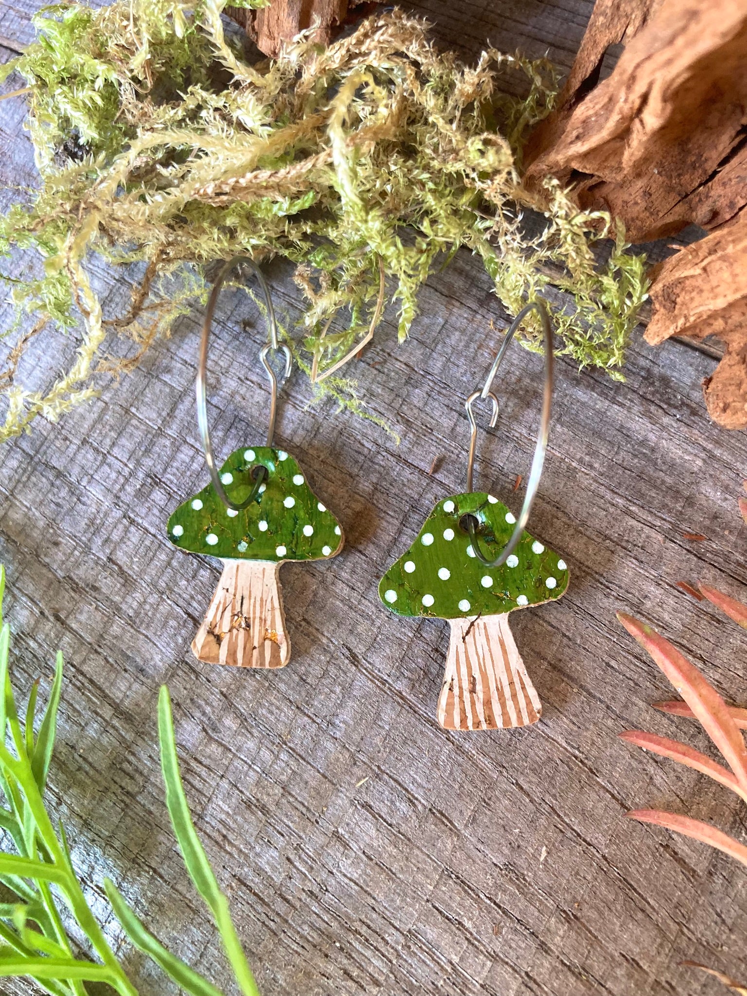 Toadstool-shaped earrings with green caps and white spots on a wooden surface with moss and leaves.
