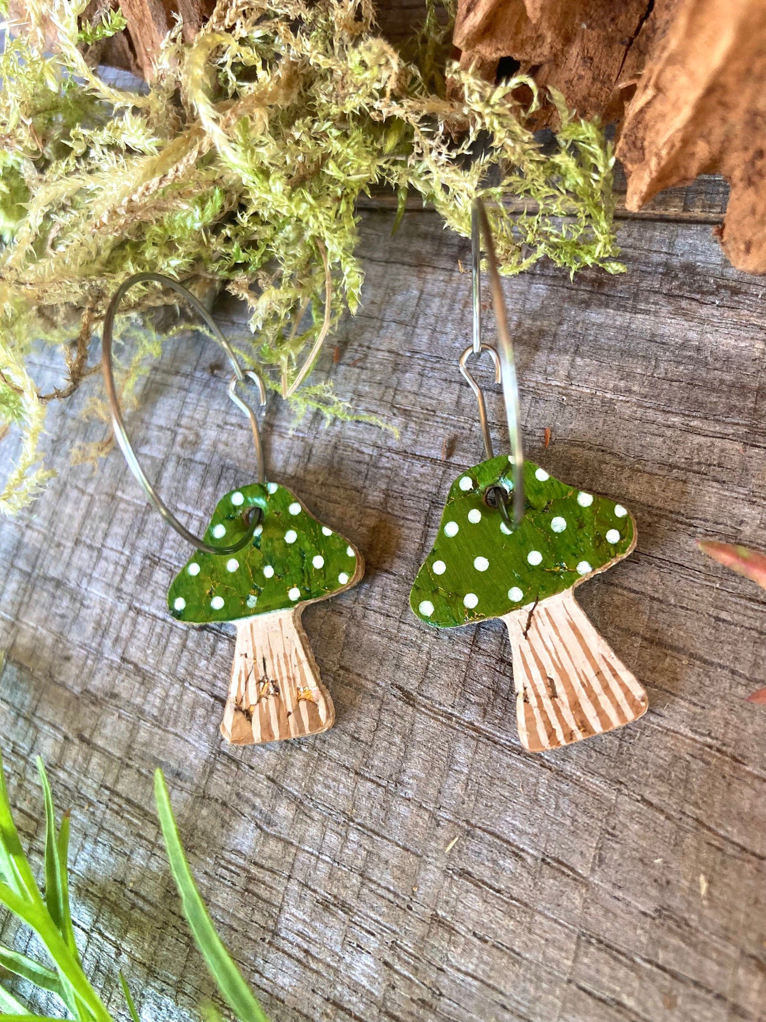 Pair of earrings shaped like green mushrooms with white spots on a wooden surface.