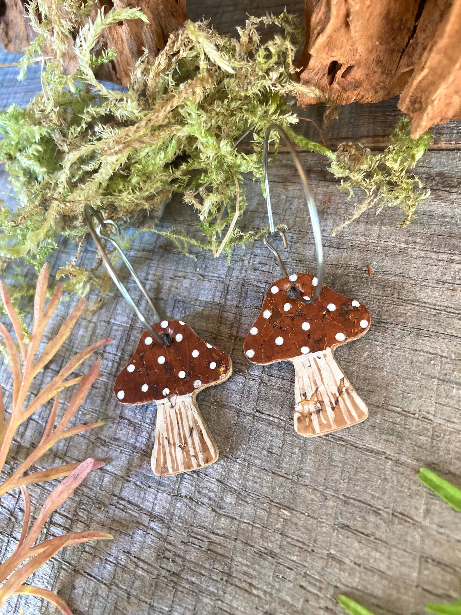 Brown toadstool shaped earrings crafted from cork with polka dot patterns on a wooden surface with moss.