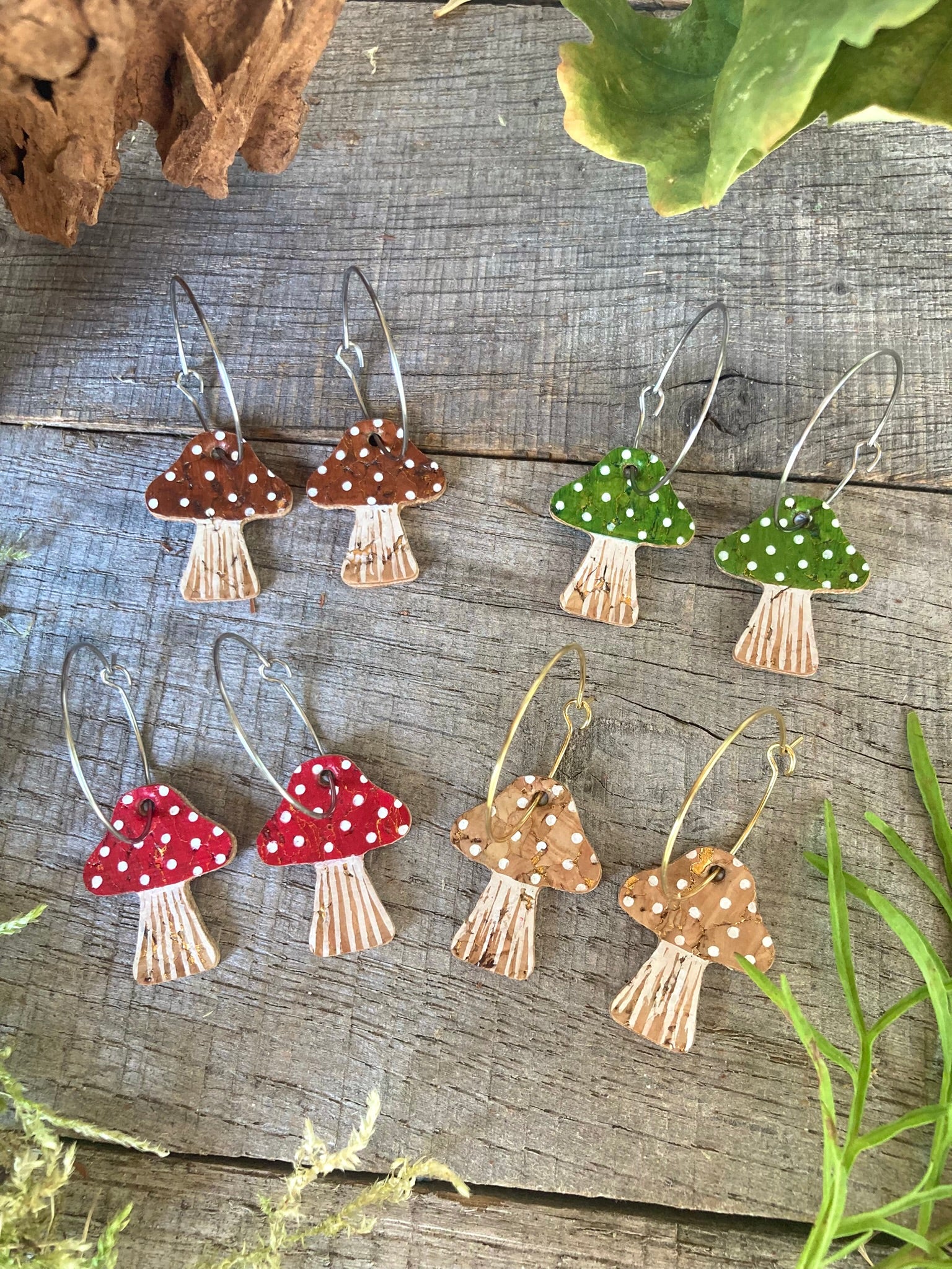 Set of mushroom-shaped earrings in 4 colourways (red, brown, green and beige with white polka dots) on a wooden surface with greenery.