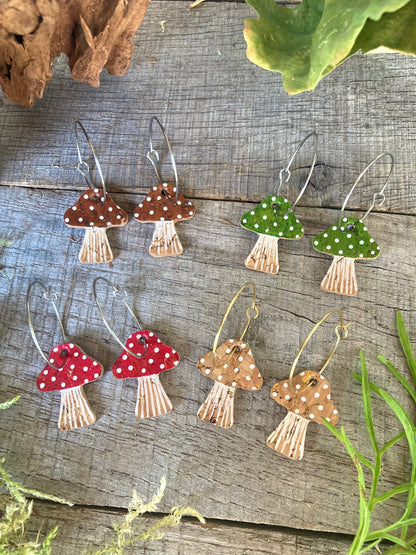 Set of mushroom-shaped earrings in 4 colourways (red, brown, green and beige with white polka dots) on a wooden surface with greenery.