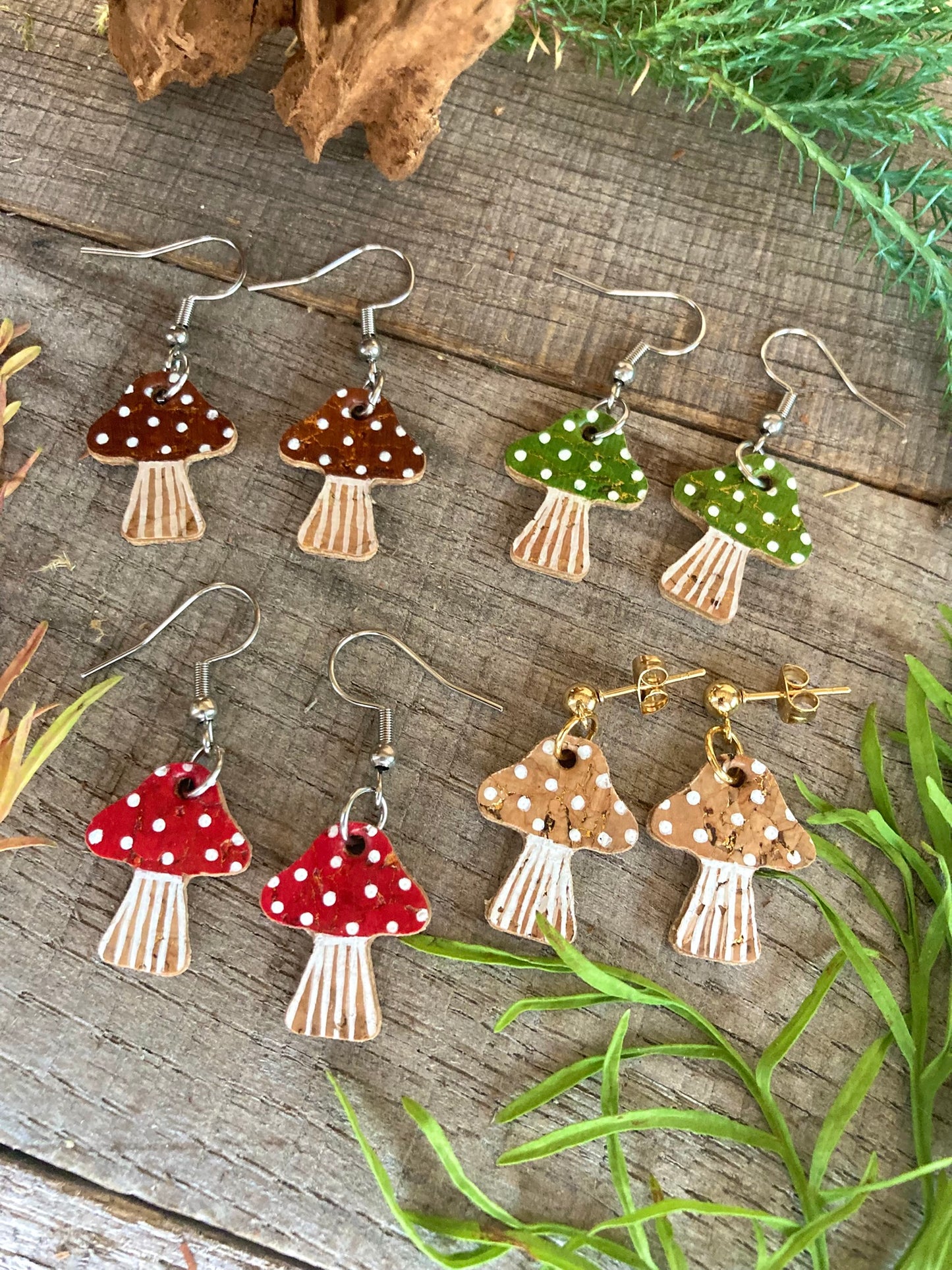 Mushroom-shaped earrings with hook or ball stud fastenings in red, green, brown and beige, on a wooden surface with greenery