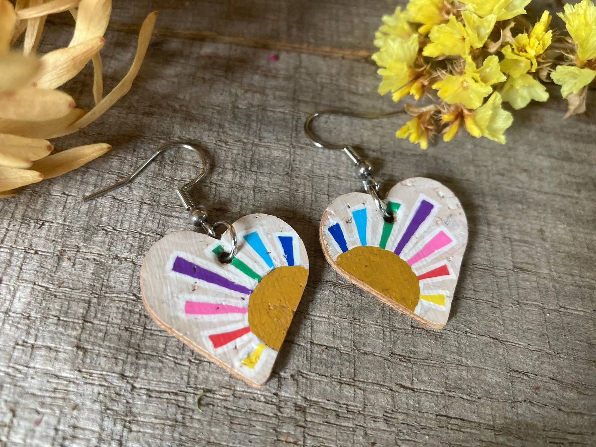 Heart-shaped earrings with colourful sun design with the rays in different colours on a wooden surface with flowers.