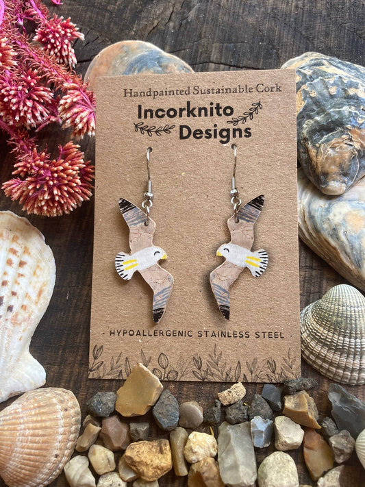 Seagull shaped earrings on a 'Incorknito Designs' card with seashells and stones in the background
