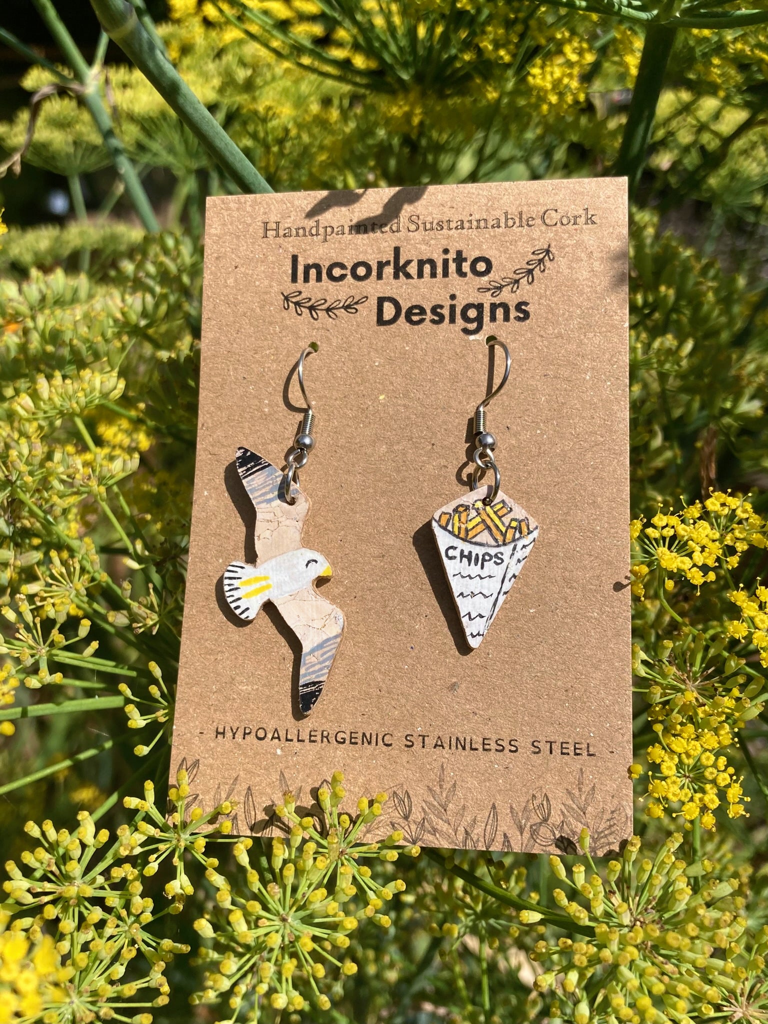 Pair of flying seagull and chips earrings on a 'Incorknito Designs' card with green foliage in the background