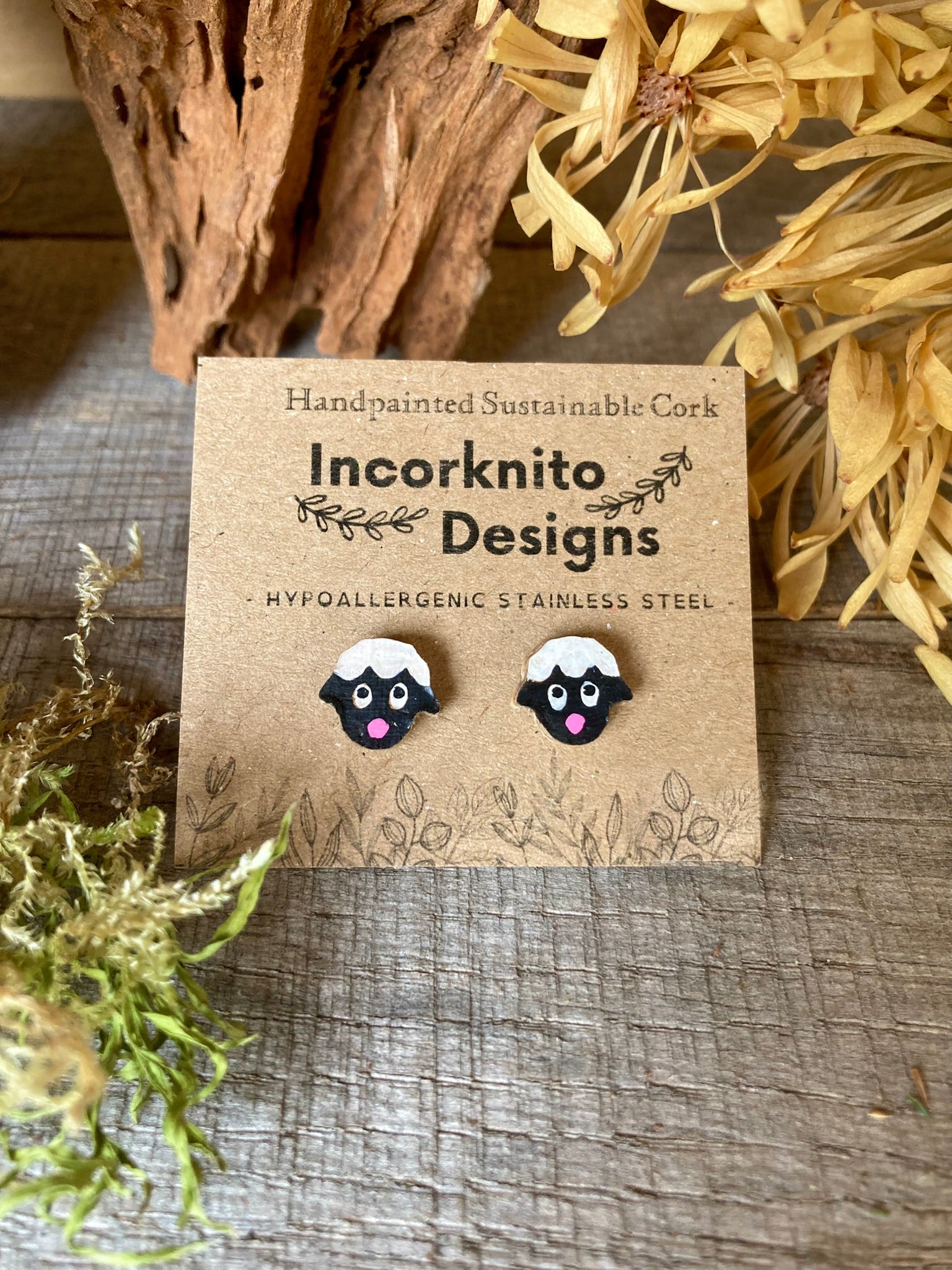 Pair of sheep-shaped earrings on a card with 'Incorknito Designs' branding, placed on a wooden surface.