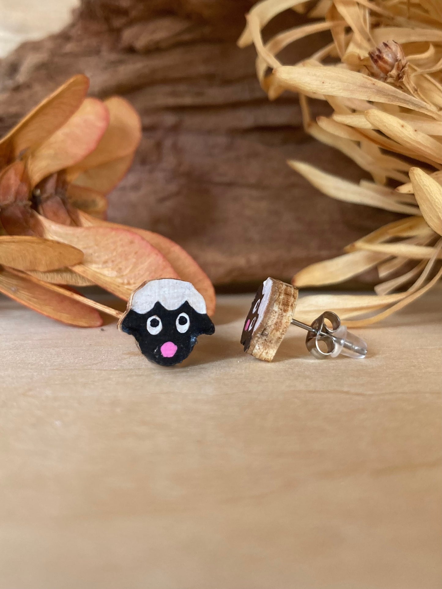 Sheep face shaped earrings on a wooden surface with dried leaves in the background