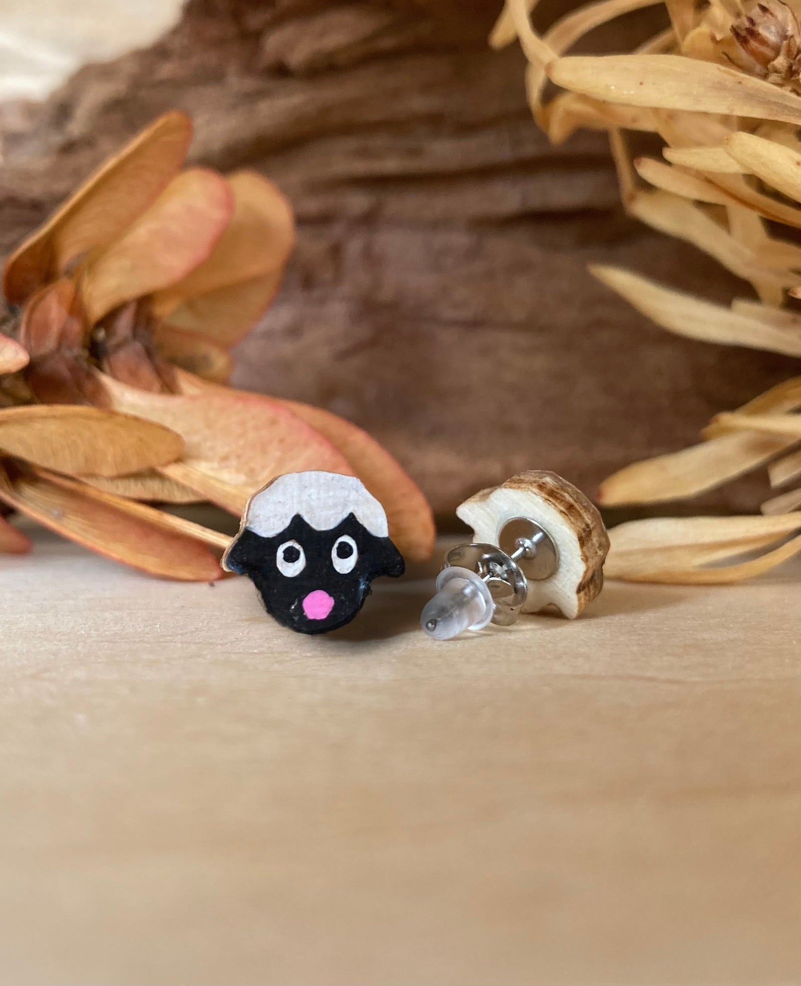 Sheep-shaped earrings on a wooden surface with dried leaves in the background