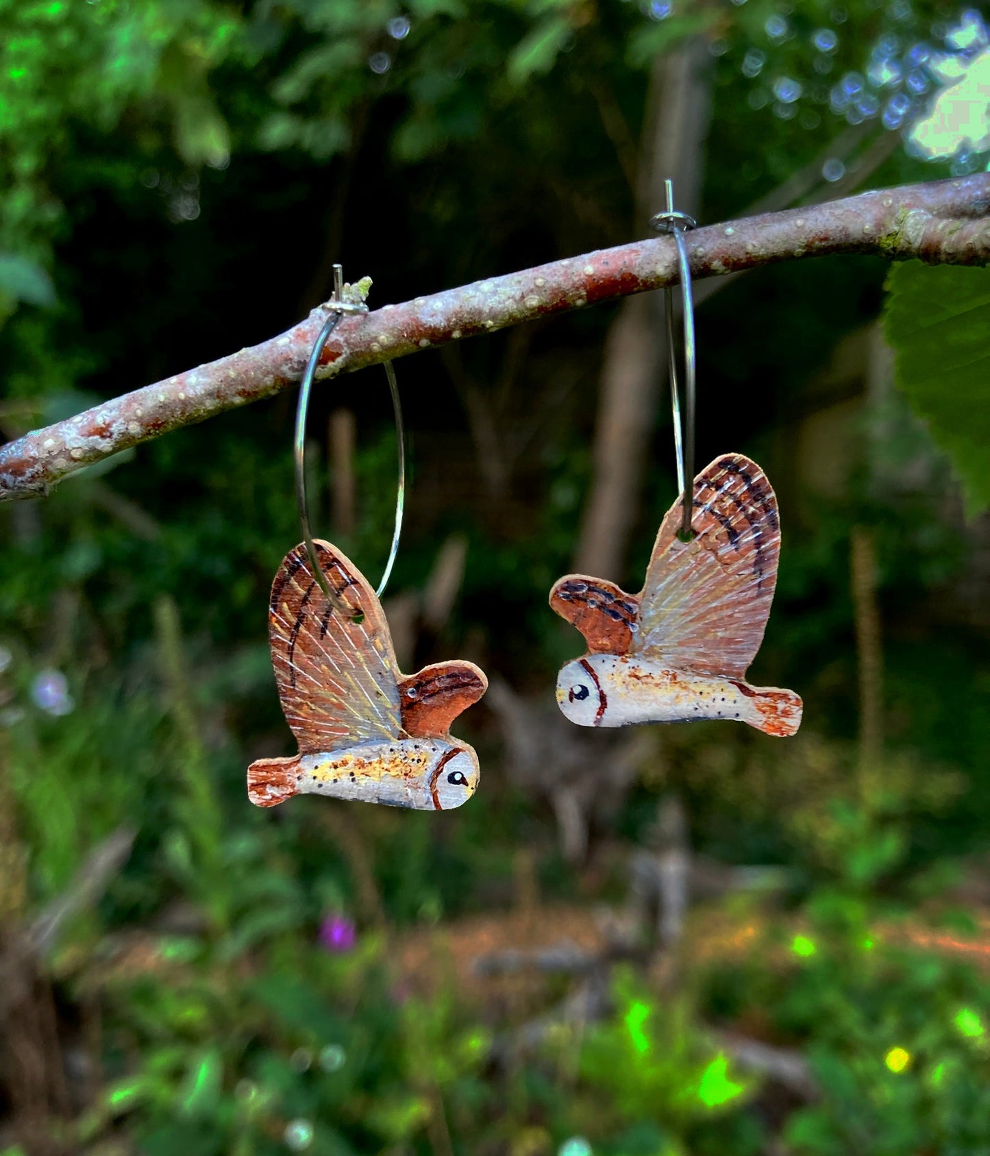 Two owl-shaped earrings hanging on a branch with a forest background