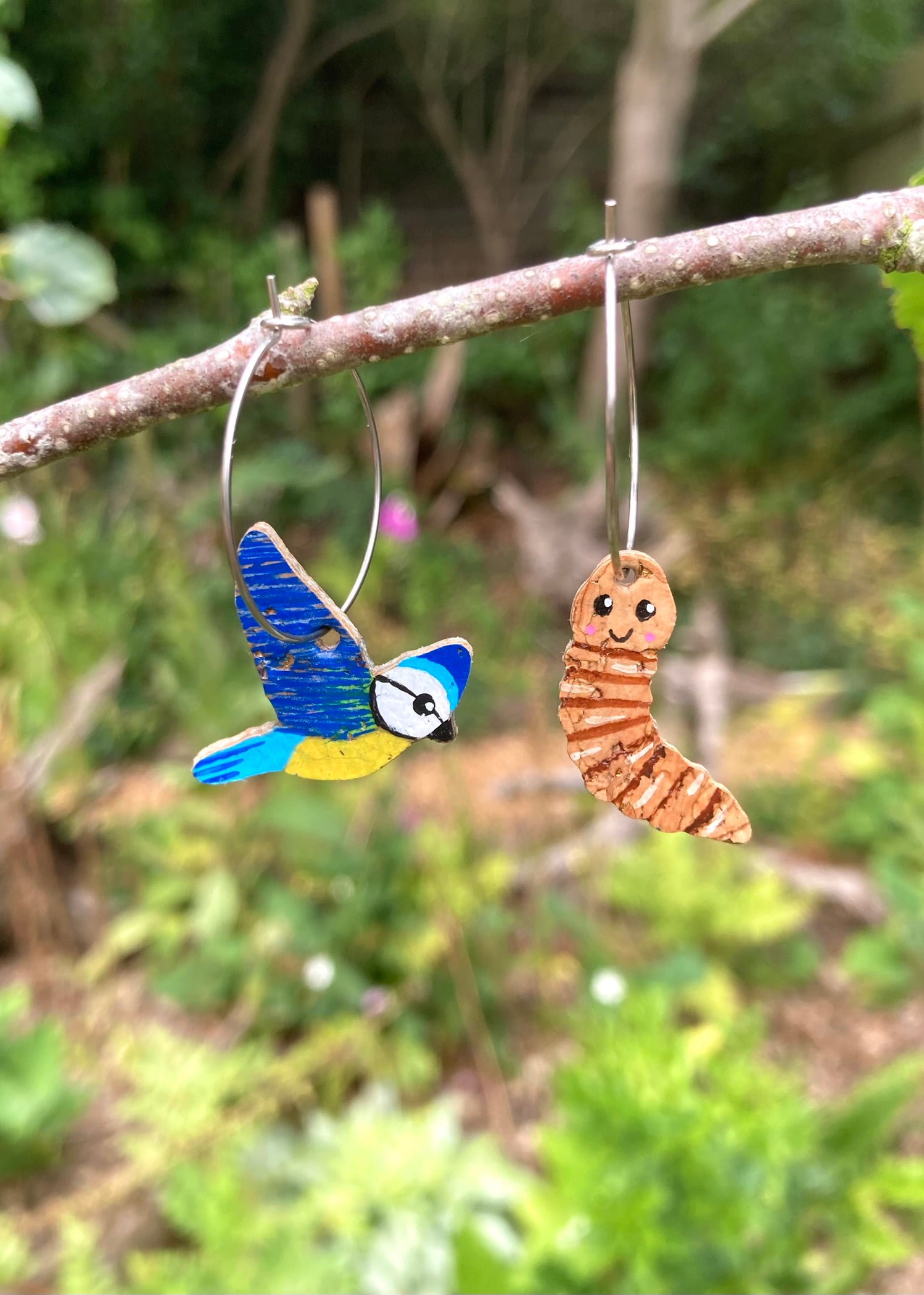 Two earrings shaped like a blue bird and a brown worm hanging on a branch with a natural background.