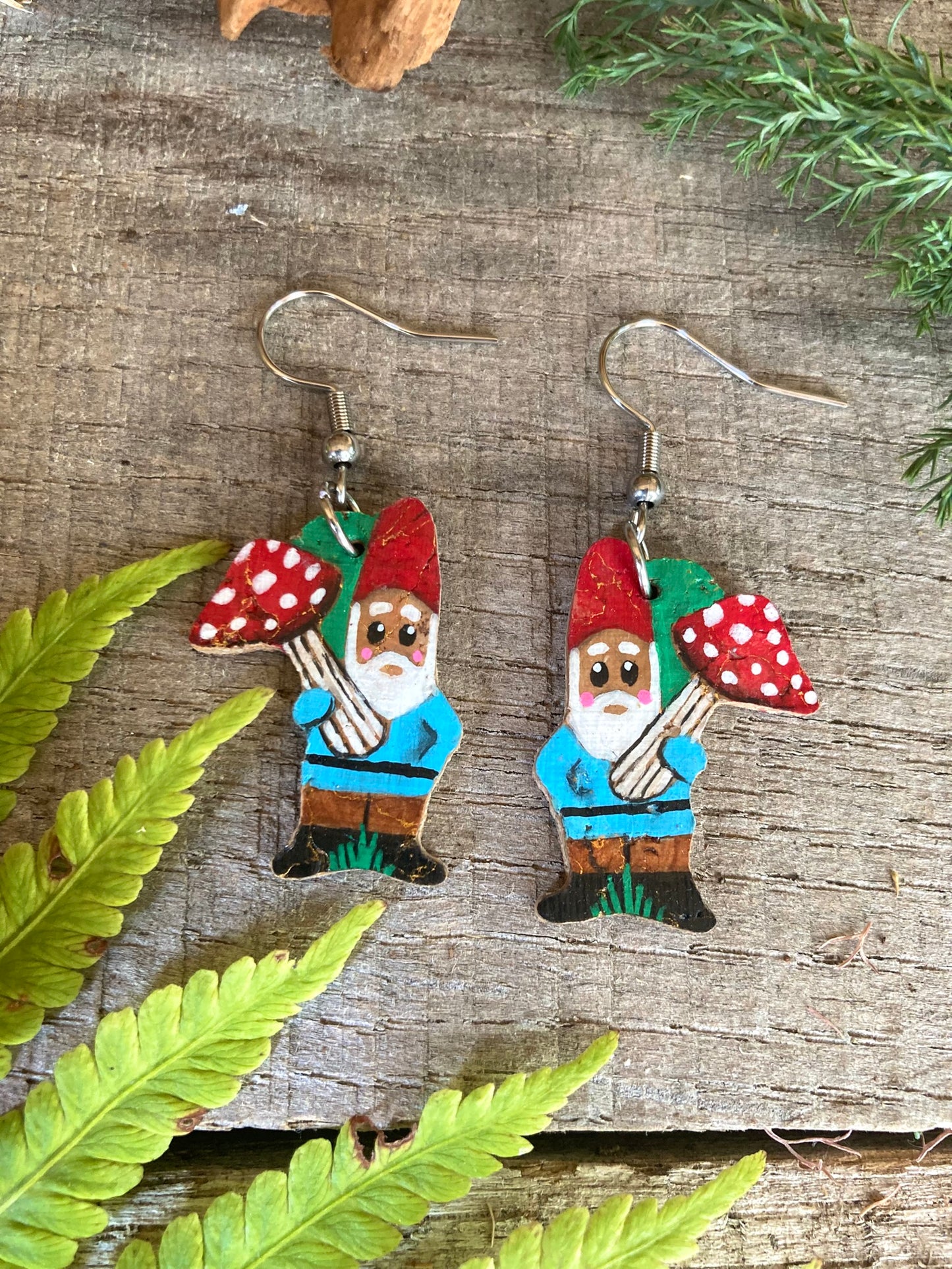 Wooden gnome earrings with red hats and mushrooms on a wooden surface with green leaves.
