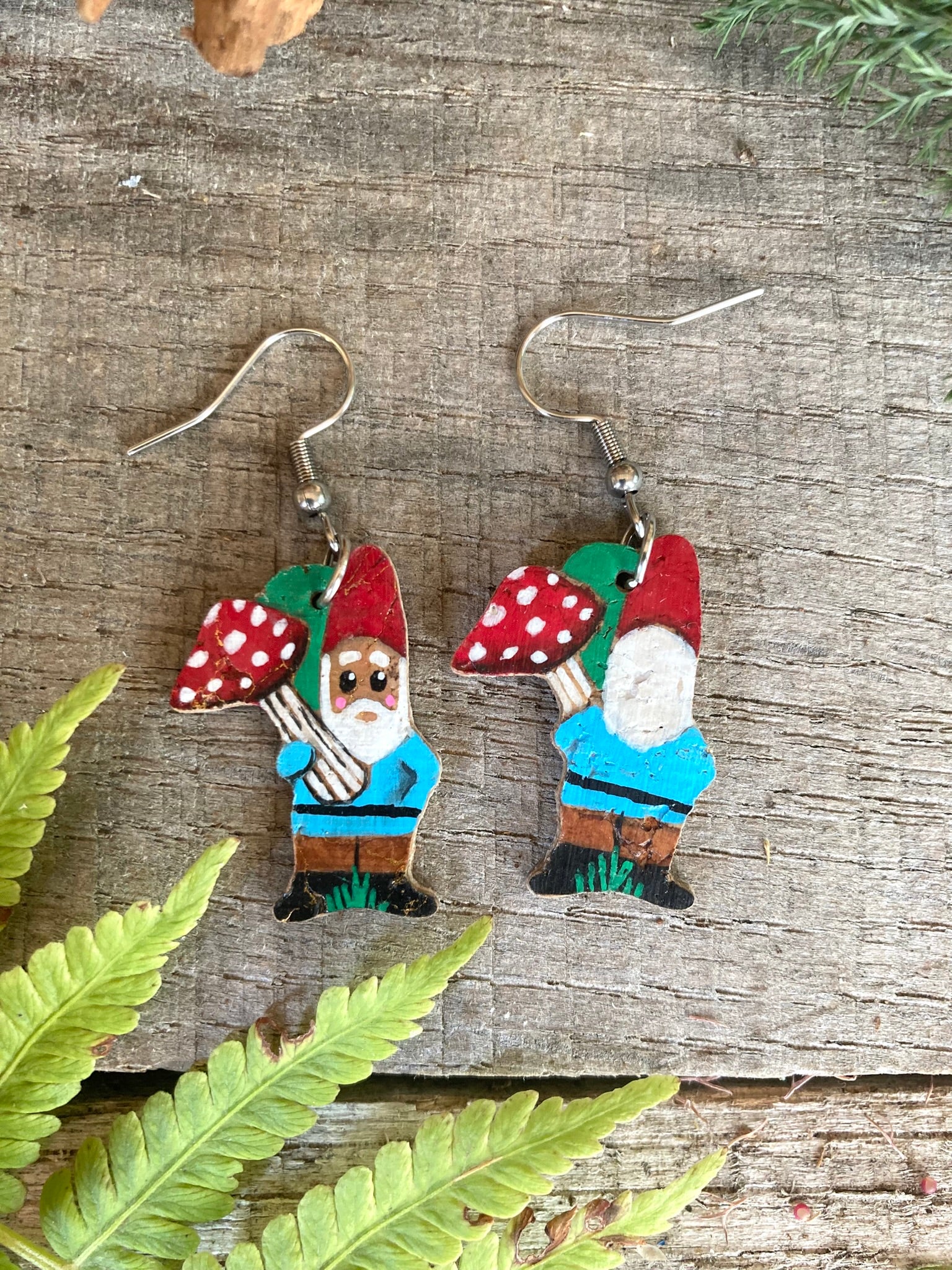 Gnome earrings with red hats holding mushrooms on a wooden surface with fern leaves.