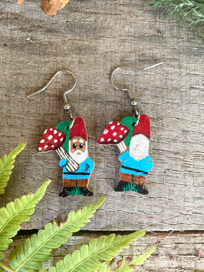 Gnome earrings with red hats holding mushrooms on a wooden surface with fern leaves.